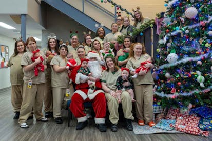 A group of female inmates in beige jumpsuits hold their babies next to a large Christmas tree, with Santa Claus in the middle.