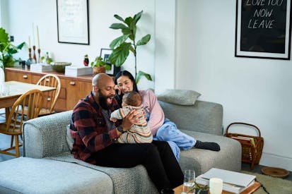 Multiracial parents sitting on sofa with baby son on black mid adult father's lap and Chinese mature mother smiling - stock photo