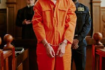 Photo: AnnaStills/Getty Images Caucasian young adult man standing in courtroom wearing handcuffs and orange jumpsuit with hands in front, police officer and judge visible in background during legal proceedings