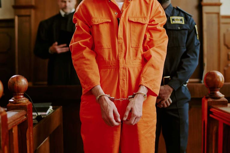 Caucasian young adult man standing in courtroom wearing handcuffs and orange jumpsuit with hands in front, police officer and judge visible in background during legal proceedings