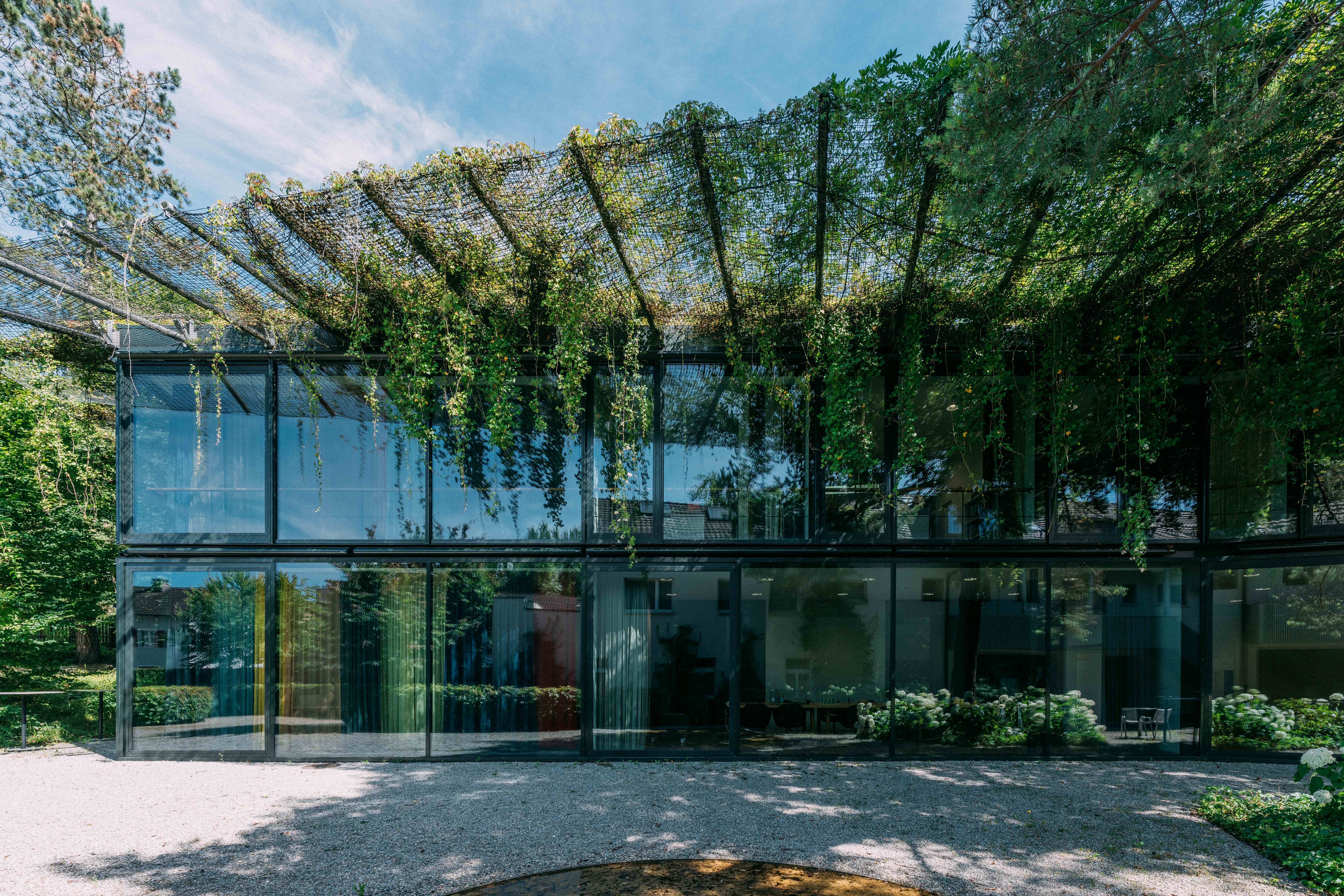 Moderno edificio de cristal rodeado de plantas y flores blancas, con un cielo despejado arriba.