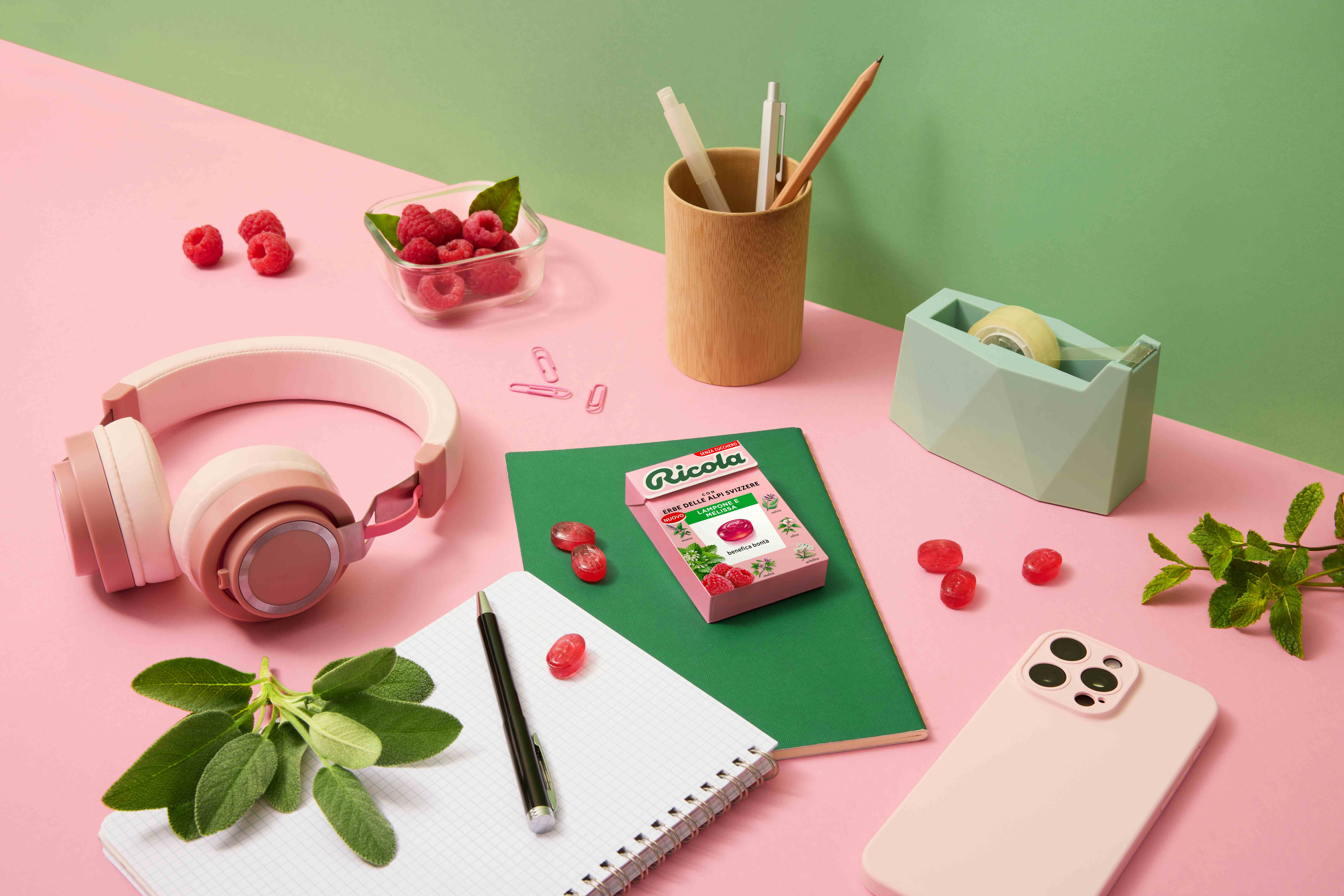 Uno spazio di lavoro colorato con cuffie rosa, un taccuino, una cartellina verde, lamponi e una confezione di Ricola Lampone Melissa su uno sfondo rosa e verde.