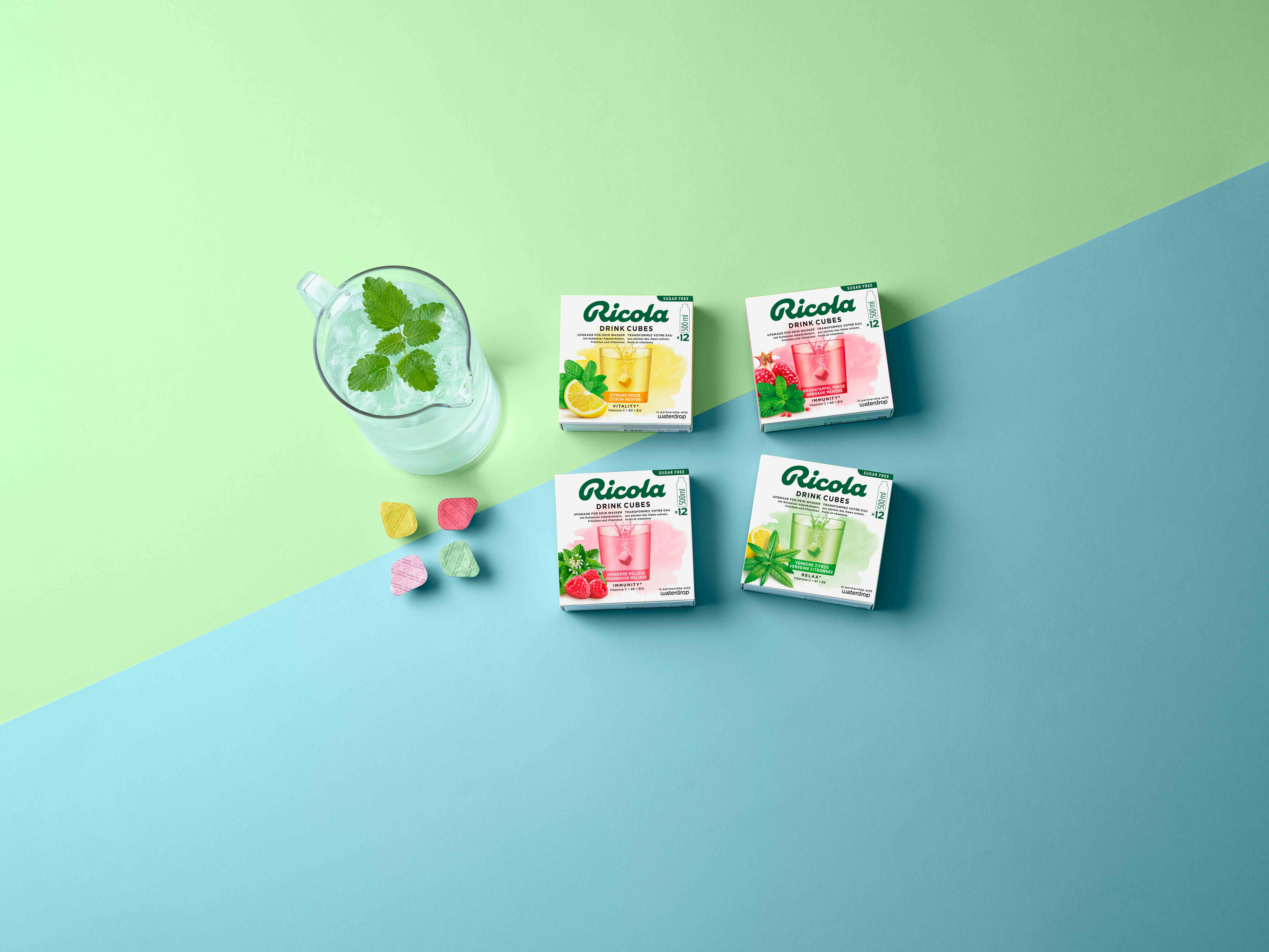 Tous les cubes Ricola Drink Cubes en un coup d'œil, avec un verre de boisson à la menthe sur un fond bleu-vert doux.