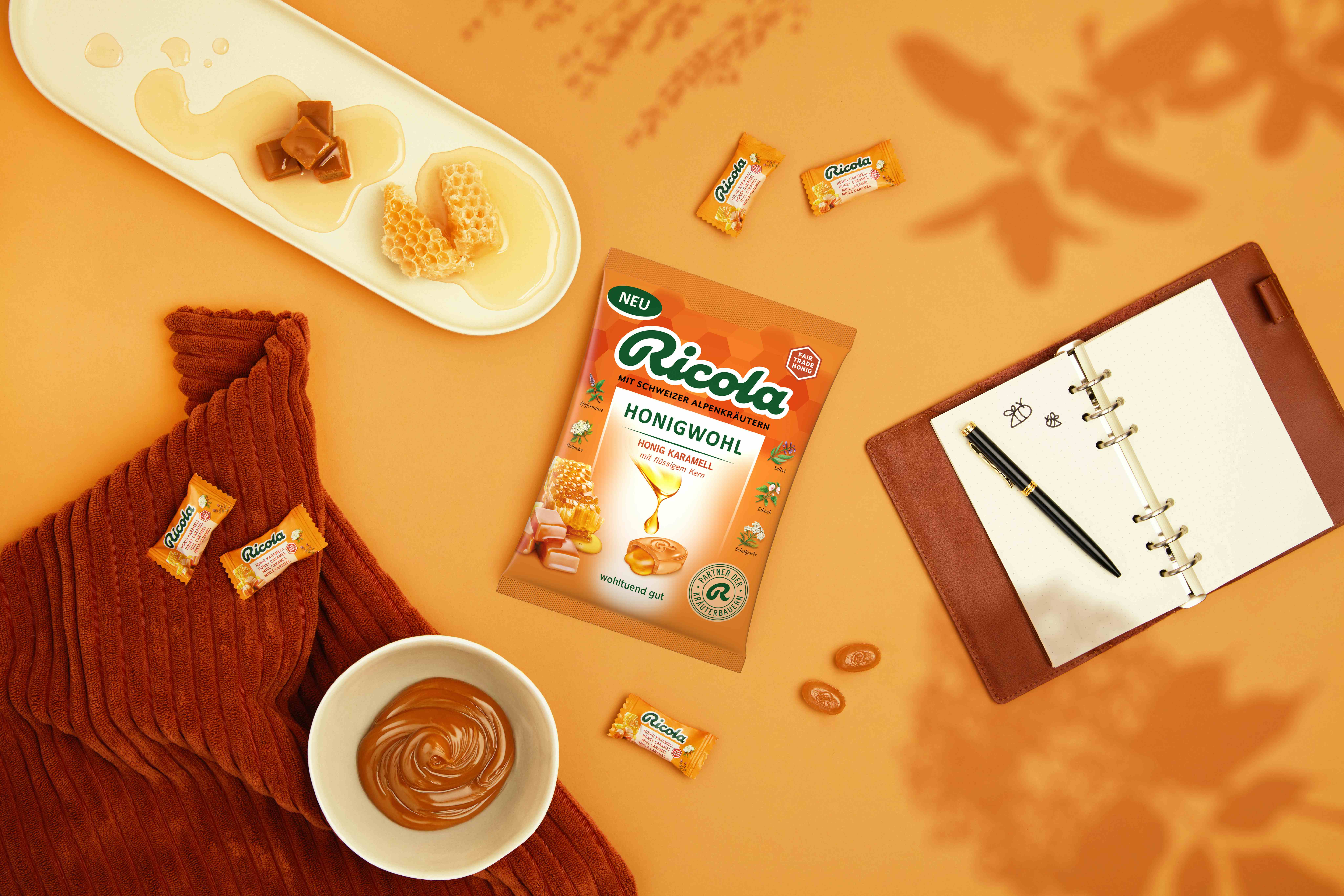 Ricola-Kräuterbonbons mit Honiggeschmack, Karamell, Waben, ein Notizbuch und ein Stift auf orangefarbenem Hintergrund.