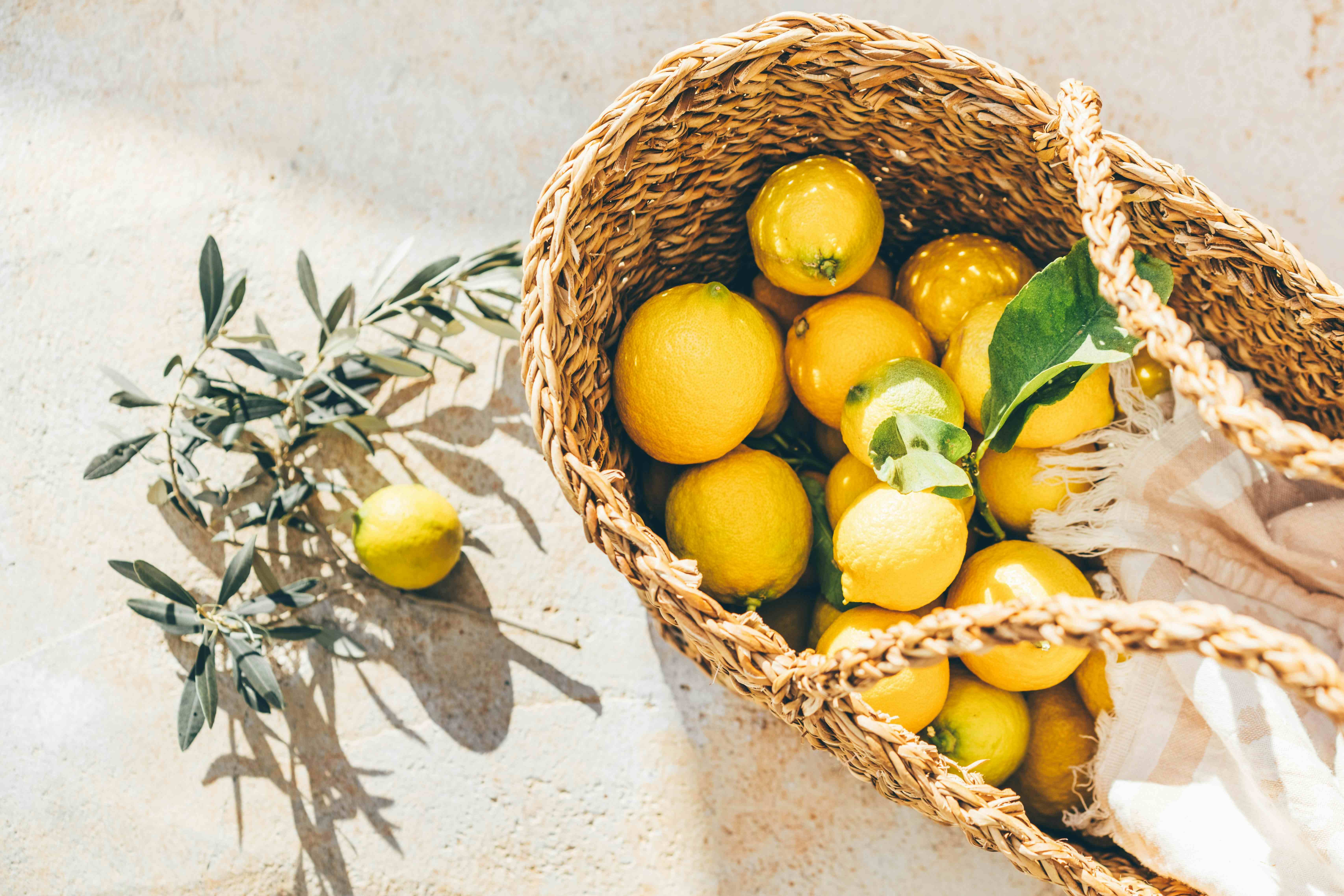 Top-down view of bright yellow lemons in a rustic straw basket on a stone floor with olive branches and sunlight.