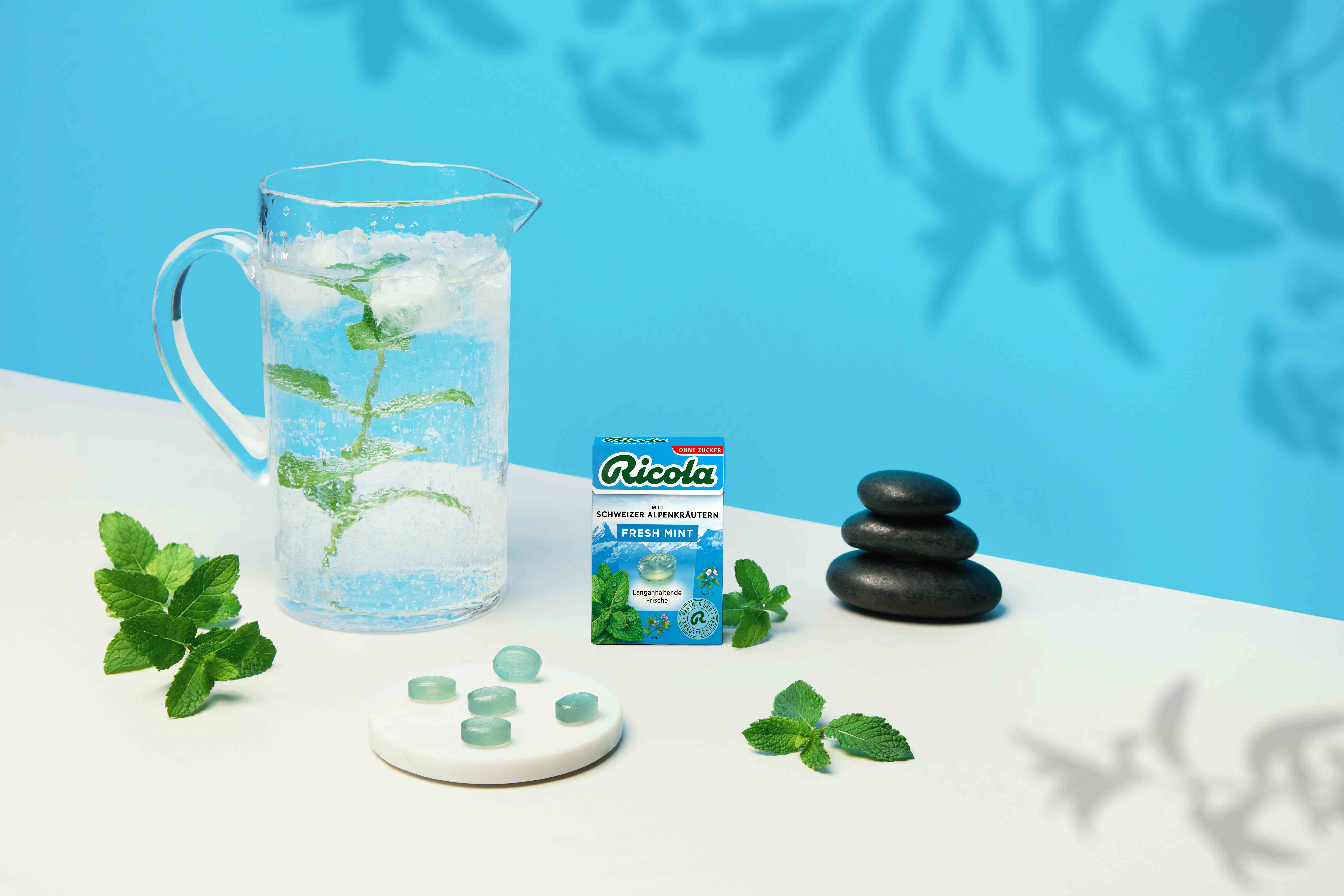 Confezione di Ricola Fresh Mint accanto a una caraffa d'acqua ghiacciata, foglie di menta e pietre nere levigate.