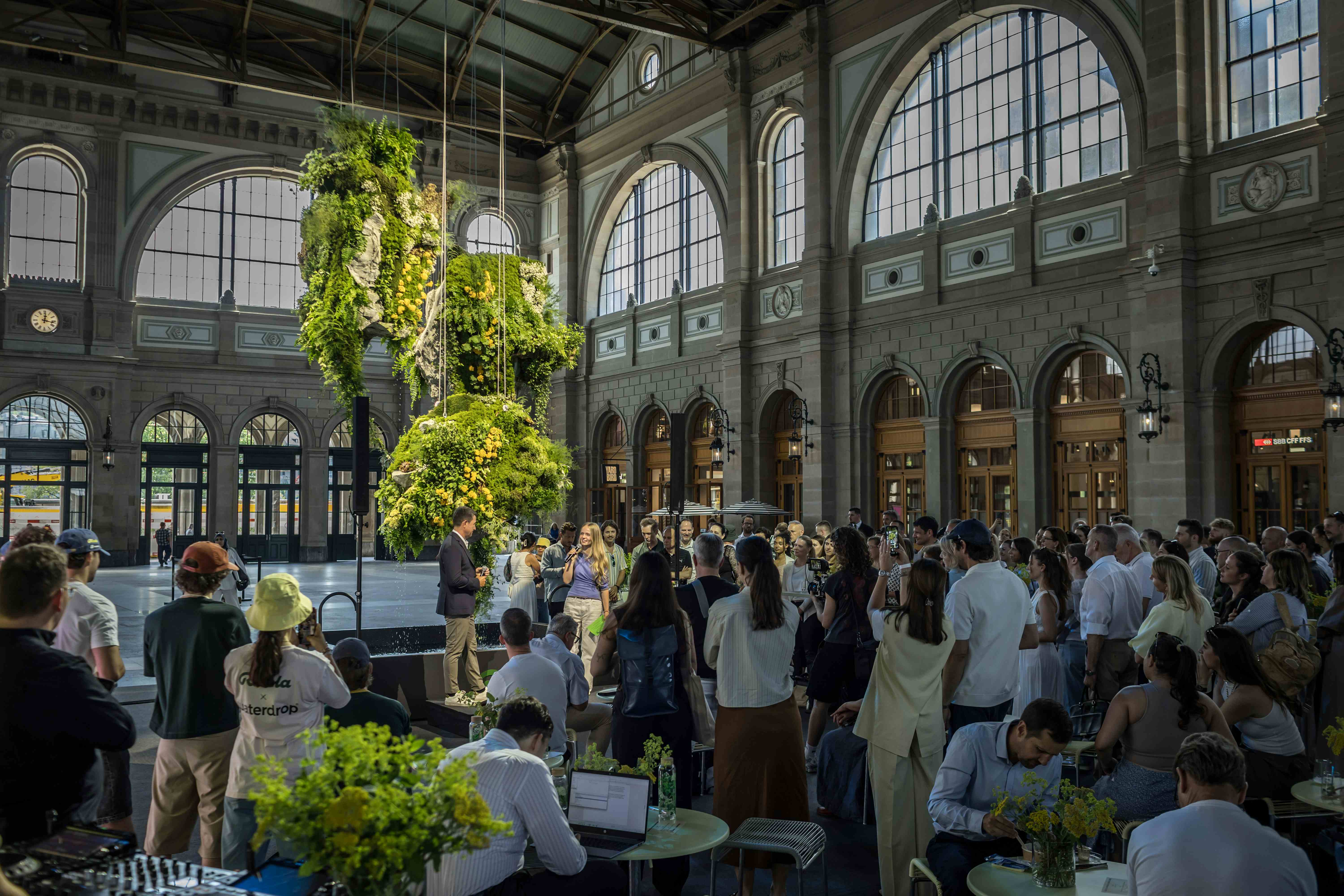 Crowd at the Ricola Drink Cubes launch event at Zurich HB, featuring a large suspended alpine herb and flower fountain installation.