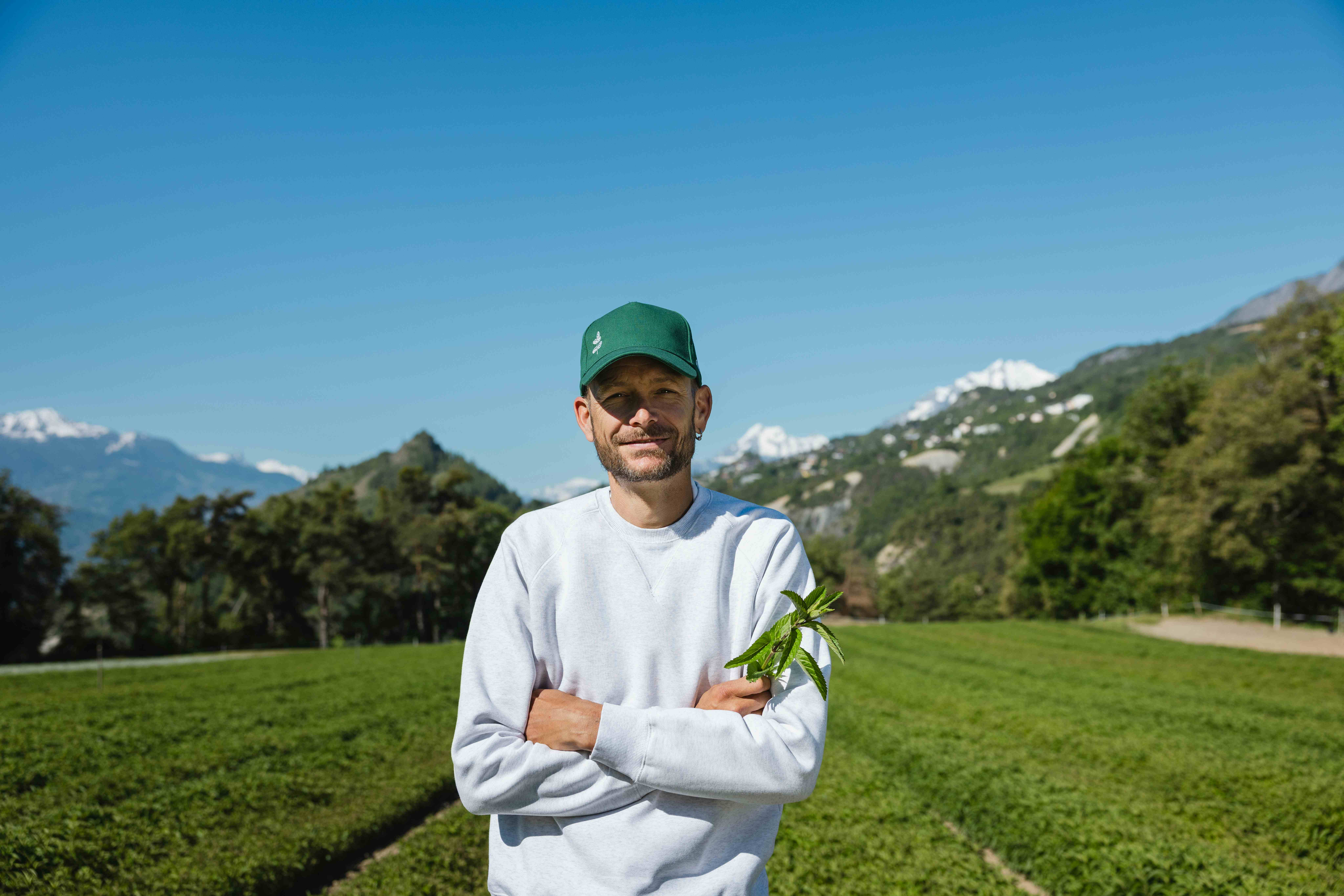A man in a white sweatshirt and green cap stands in a lush field holding a sprig of herbs, with mountains in the background.