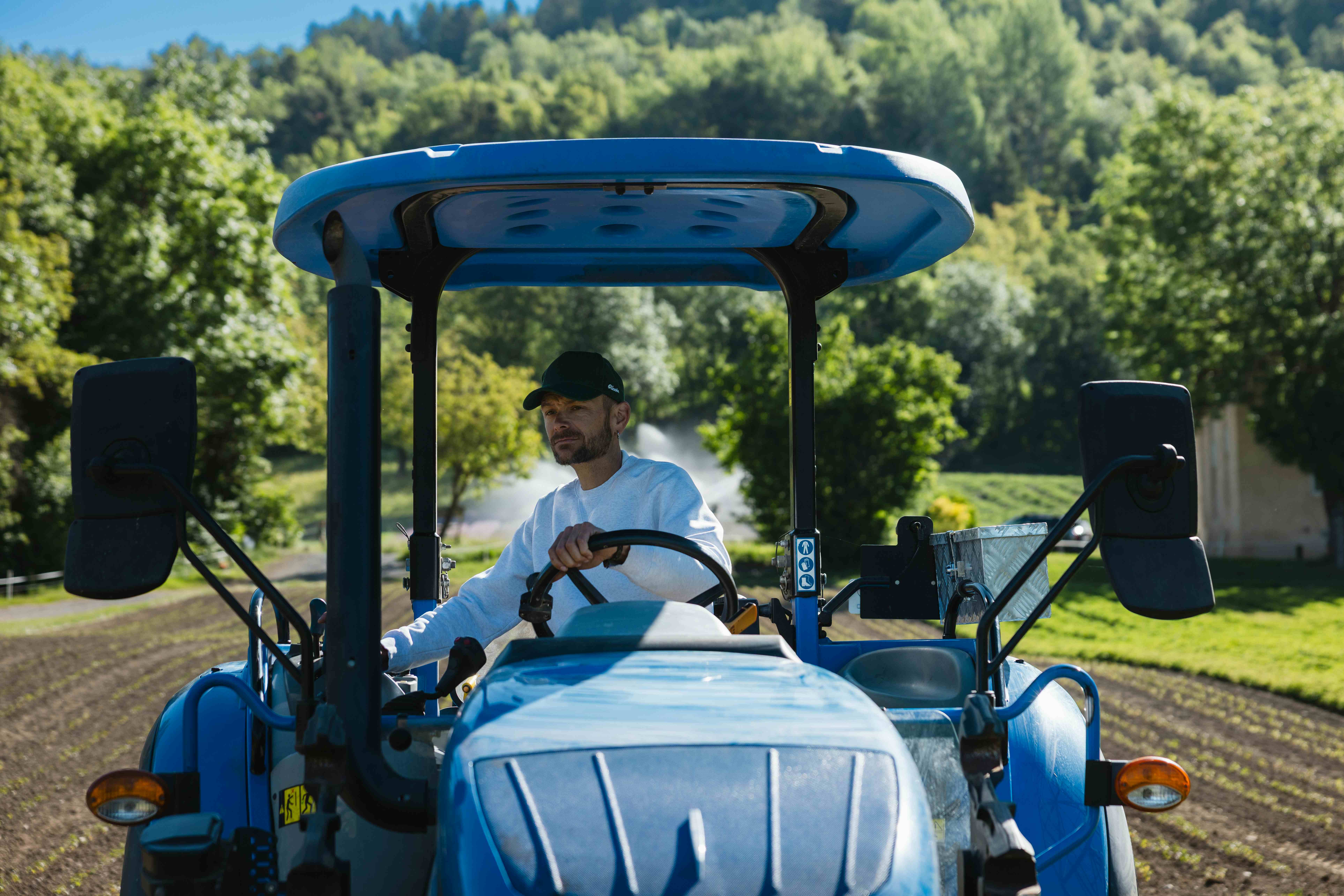 A farmer in a white shirt and green cap drives a blue tractor through a field with trees and a sprinkler in the background.