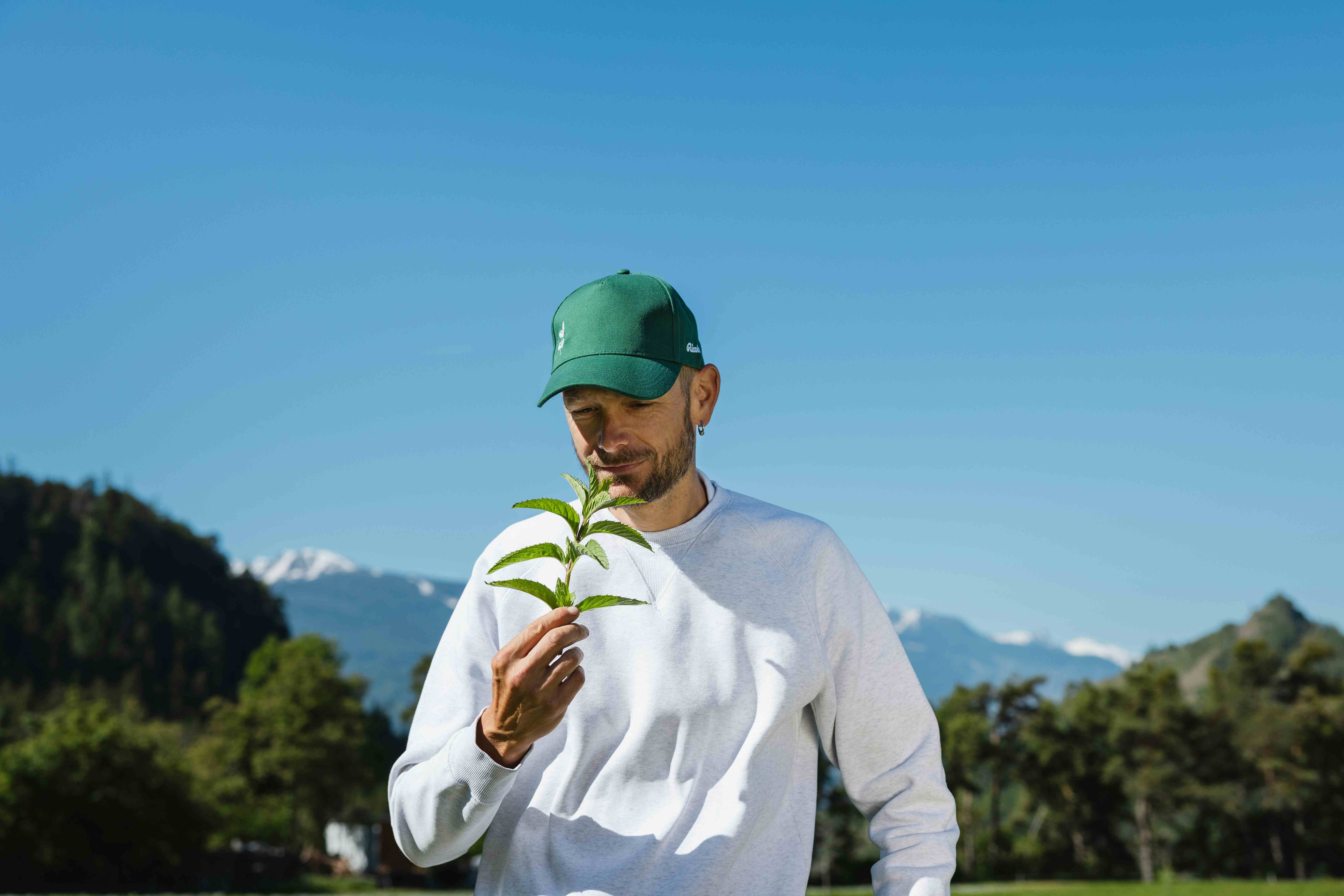 A farmer in a green cap and white shirt smells a fresh sprig of green herb outdoors against a bright blue sky and mountains.