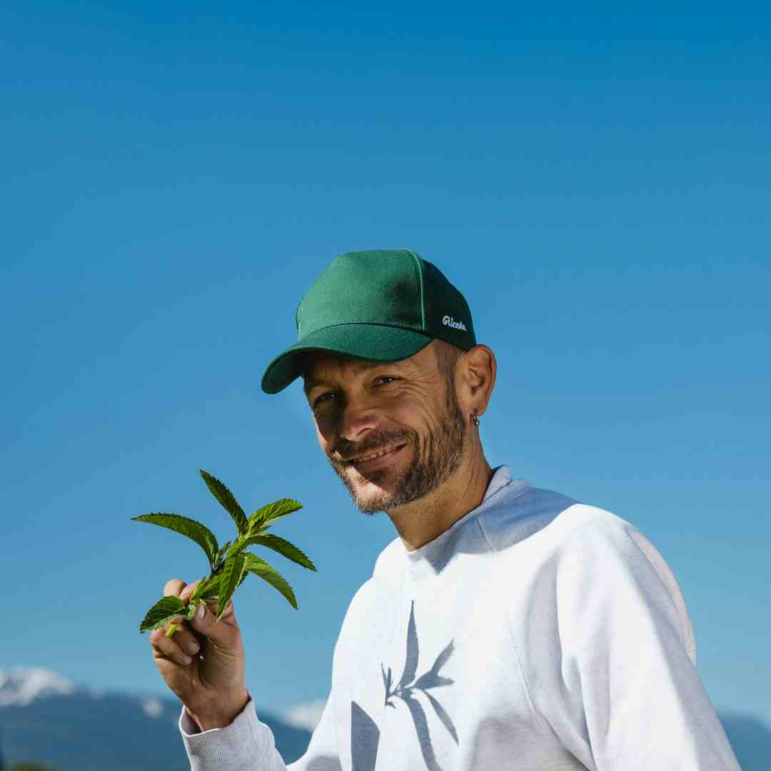 A smiling man in a green cap and white shirt holds a fresh green herb sprig against a clear blue sky.