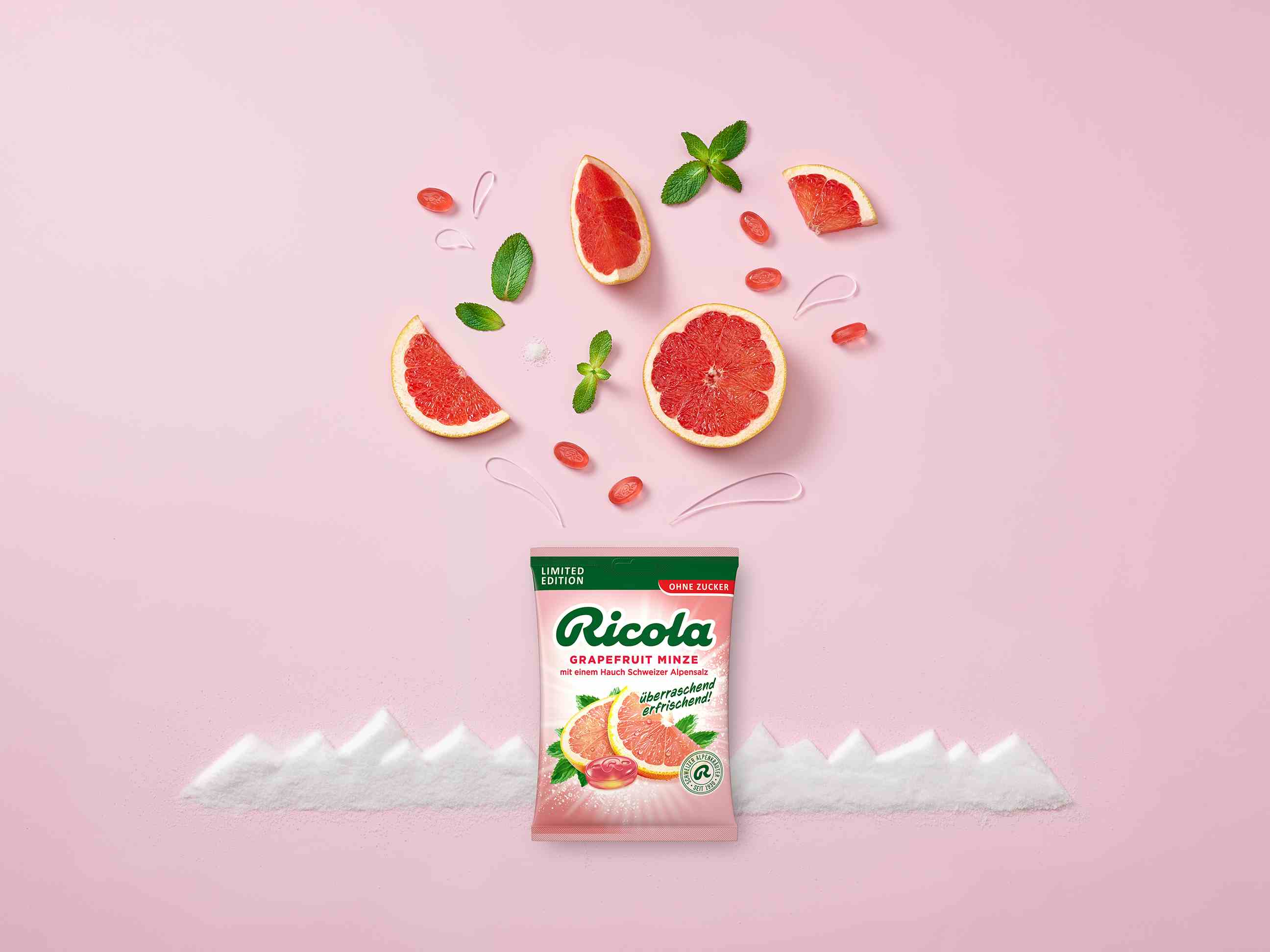 Eine Tüte Ricola zuckerfreie Bonbons der Sorte „Grapefruit Minze“ steht zentriert auf rosa Hintergrund vor einer stilisierten Gebirgskette aus weißem Salz. Über der Packung schweben Grapefruitscheiben, frische Minzblätter und rosa Bonbons in der Luft.