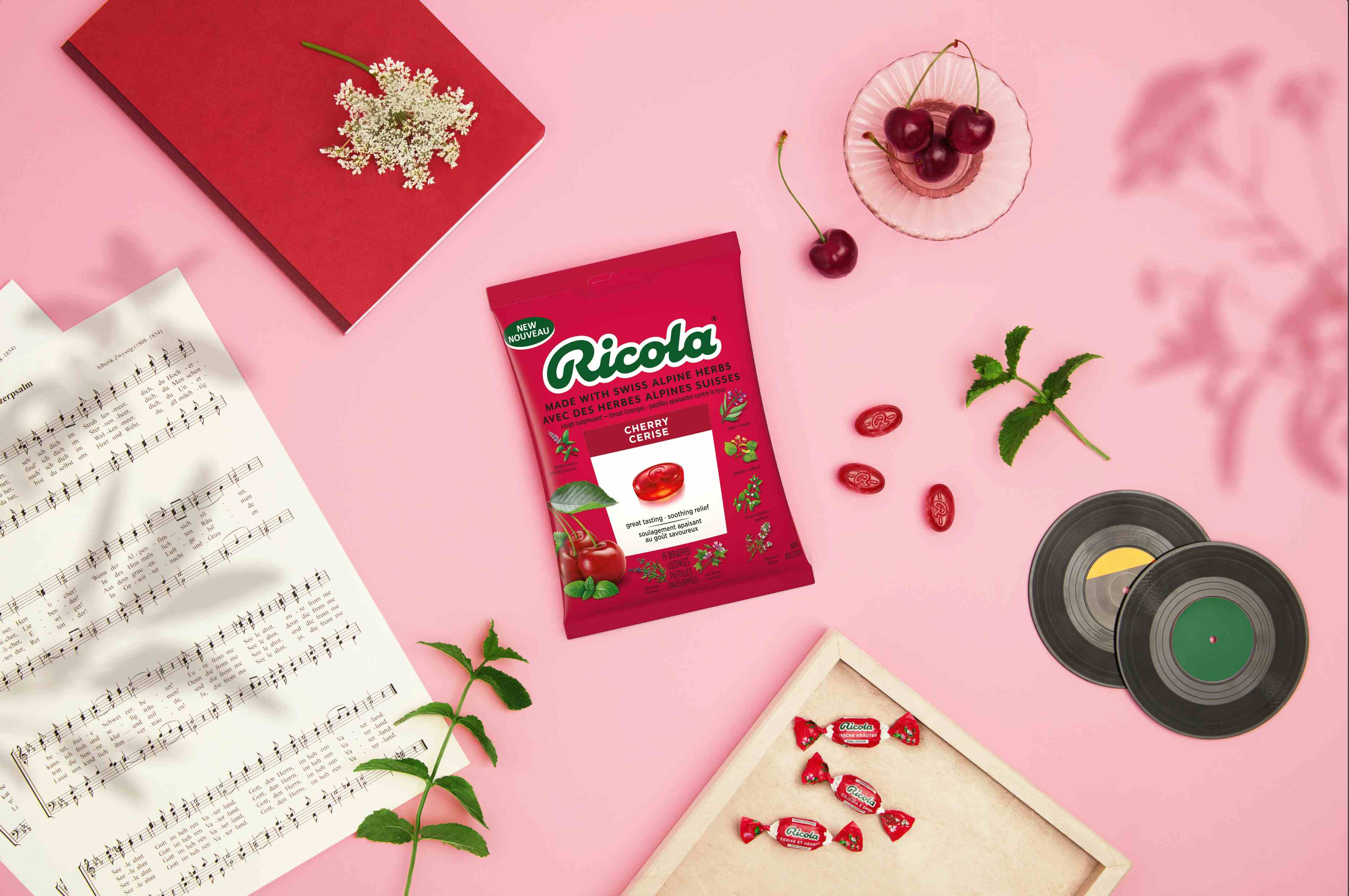 Top-down view of a Ricola Cherry Cerise bag surrounded by fresh cherries, mint sprigs, sheet music, a notebook, vinyl records, and wrapped lozenges on a pink surface.