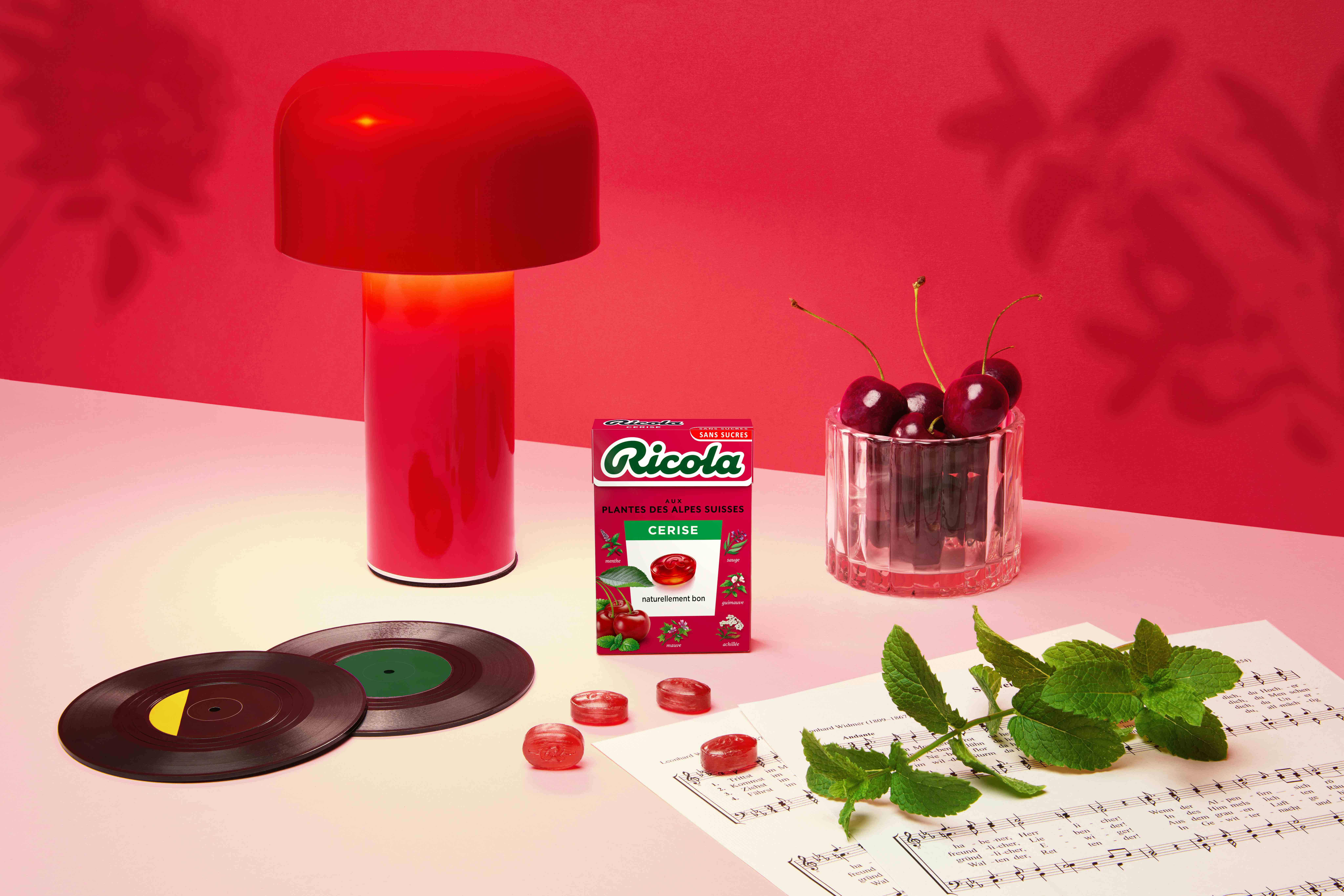 Boîte de Ricola Cerise sur une table rose près d'une lampe rouge, des cerises, des vinyles, des bonbons et de la menthe sur une partition.