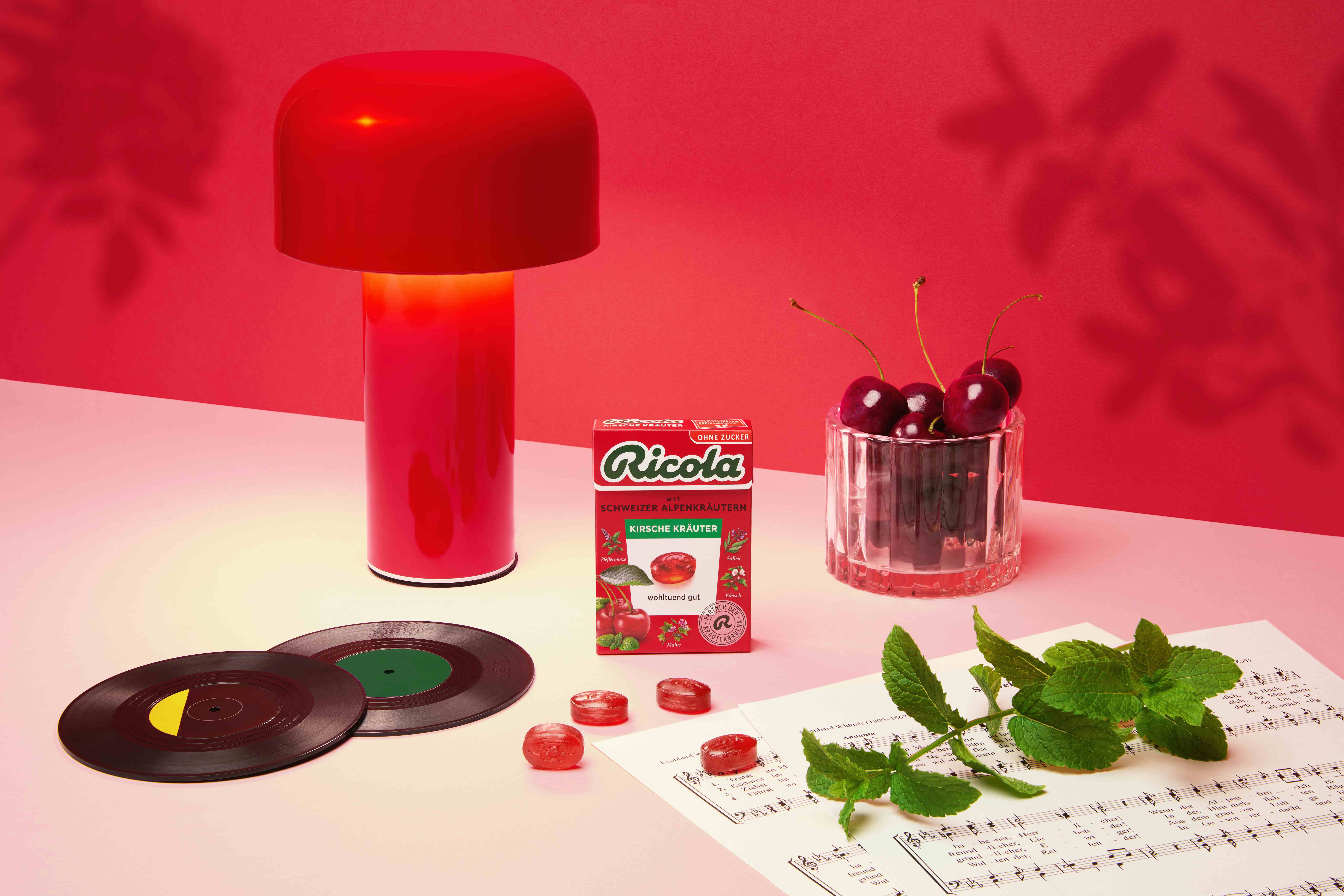 Ricola Cherry Herbs box on a pink table beside a red lamp, fresh cherries in a glass, vinyl records, loose red lozenges, and fresh mint leaves on sheet music.