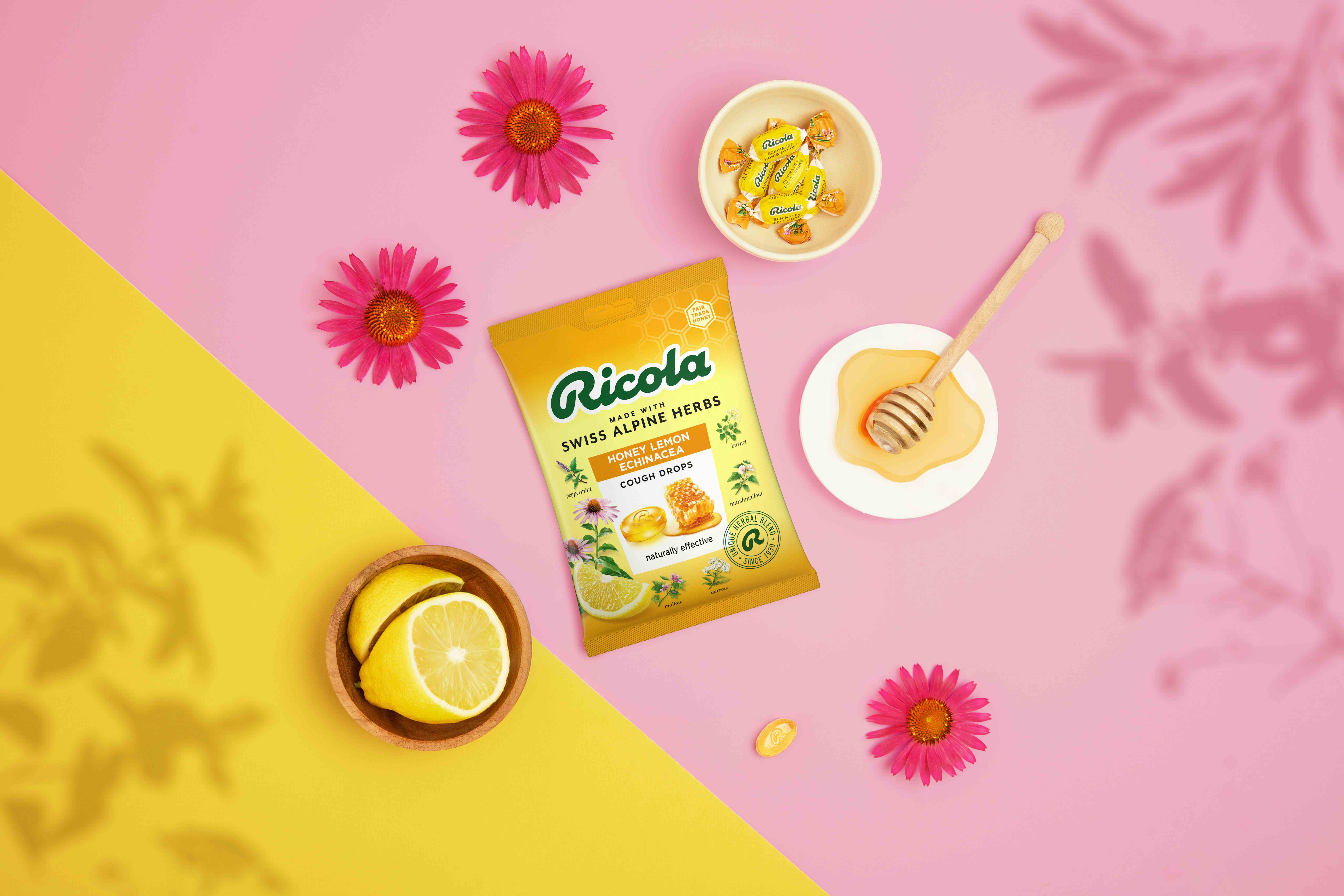 Ricola Honey Lemon Echinacea cough drops bag surrounded by lemon slices, a honey dipper, pink flowers, and wrapped candies.