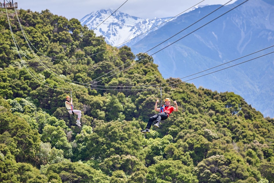 To people descend on ziplines in Kaikoura with alps behind them
