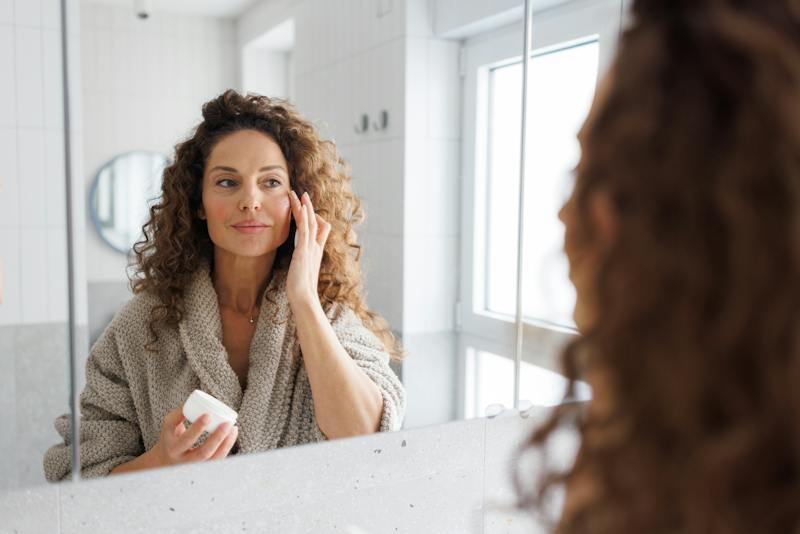 Woman looking in the mirror while applying skincare to her face.