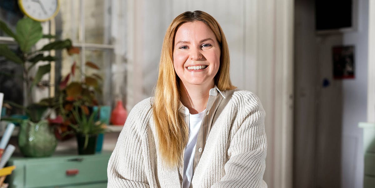 Nainen istuu kotonaan tuolissa ja katsoo kameraan hymyillen