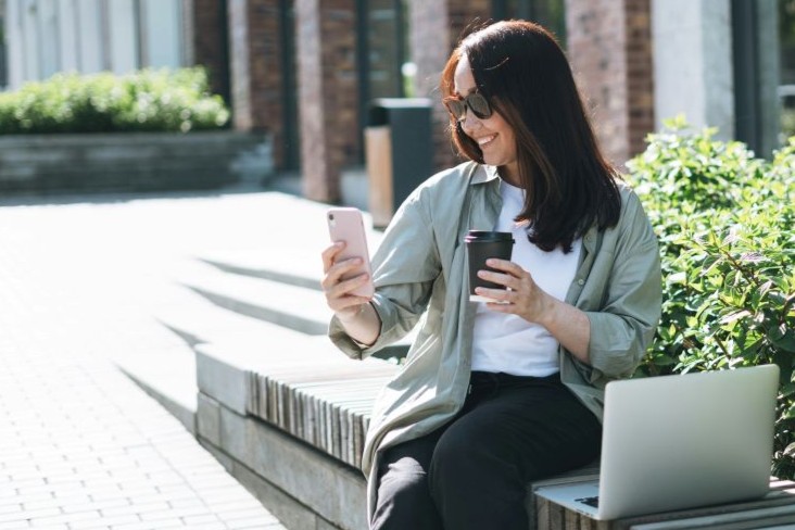 Woman outside happily on phone with coffee and laptop