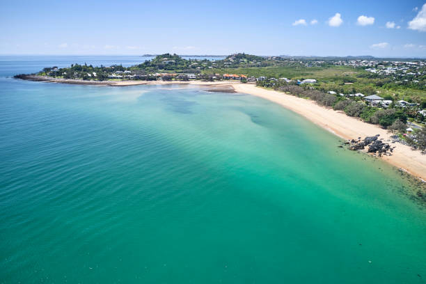 Aerial view of a serene coastline with turquoise waters gently lapping against a sandy beach. The shoreline is dotted with green vegetation and a few houses. In the distance, a small headland extends into the sea under a clear blue sky with a few white clouds.
