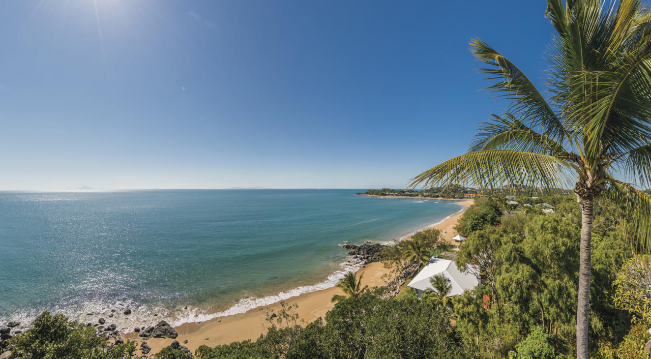 A panoramic view of a serene tropical beach with golden sand and calm blue waters. Palm trees and lush greenery frame the scene, with clear blue skies overhead. A few white tents are visible among the vegetation near the shoreline.