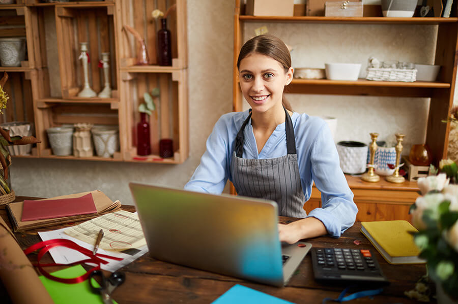 A woman wearing a light blue shirt and striped apron sits at a wooden desk in a cozy workshop. She smiles at the camera with a laptop in front of her. The background features shelves with various decorative items and supplies, including vases and books.