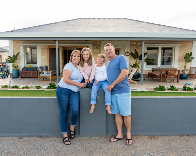 A family standing in front of a house with plants, chairs and tables on the background.