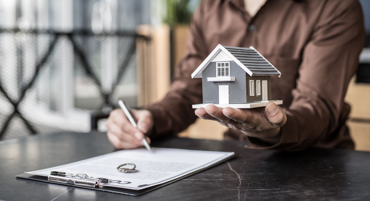 A person holding a pen on their right hand and a house on their left hand, with a house key beside.