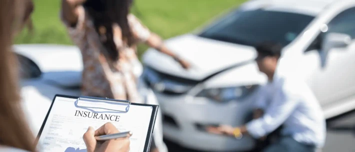 Someone holding a clipboard while writing on the insurance form with 2 vehicles on the background.