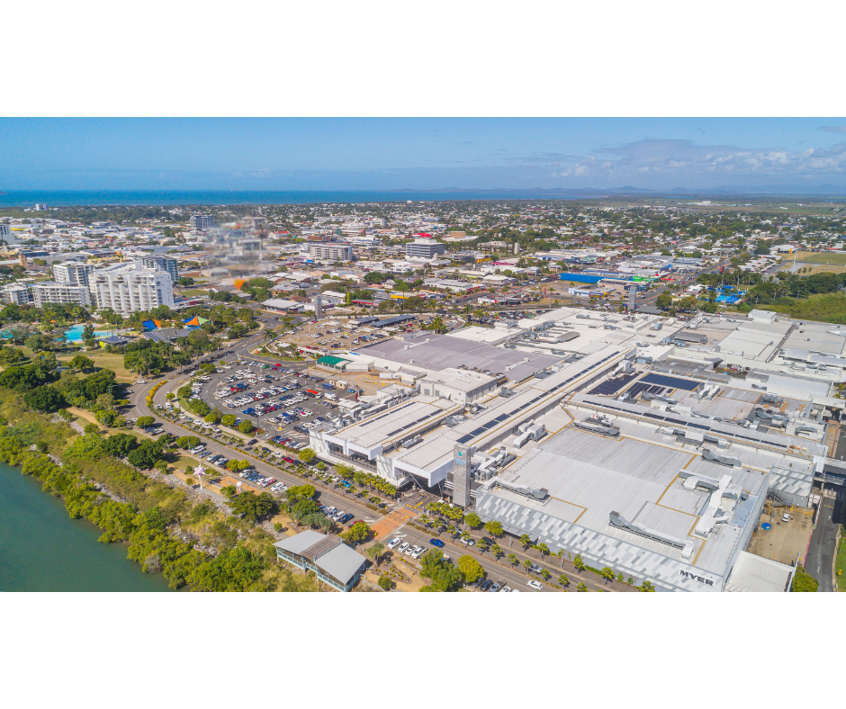 An aerial photograph showing Mackay city in Queensland, Australia, with Caneland Central shopping centre in the foreground, surrounded by parking lots, roads, and greenery, and the coastline visible in the distance under a clear blue sky.
