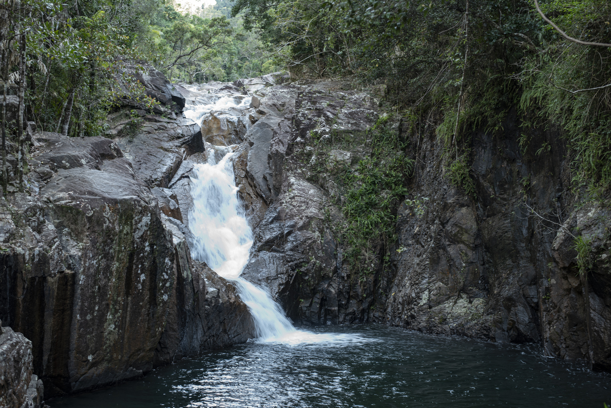 A cascading waterfall flowing over rugged rock formations into a deep pool, surrounded by lush green rainforest vegetation at Finch Hatton Gorge in Queensland, Australia.