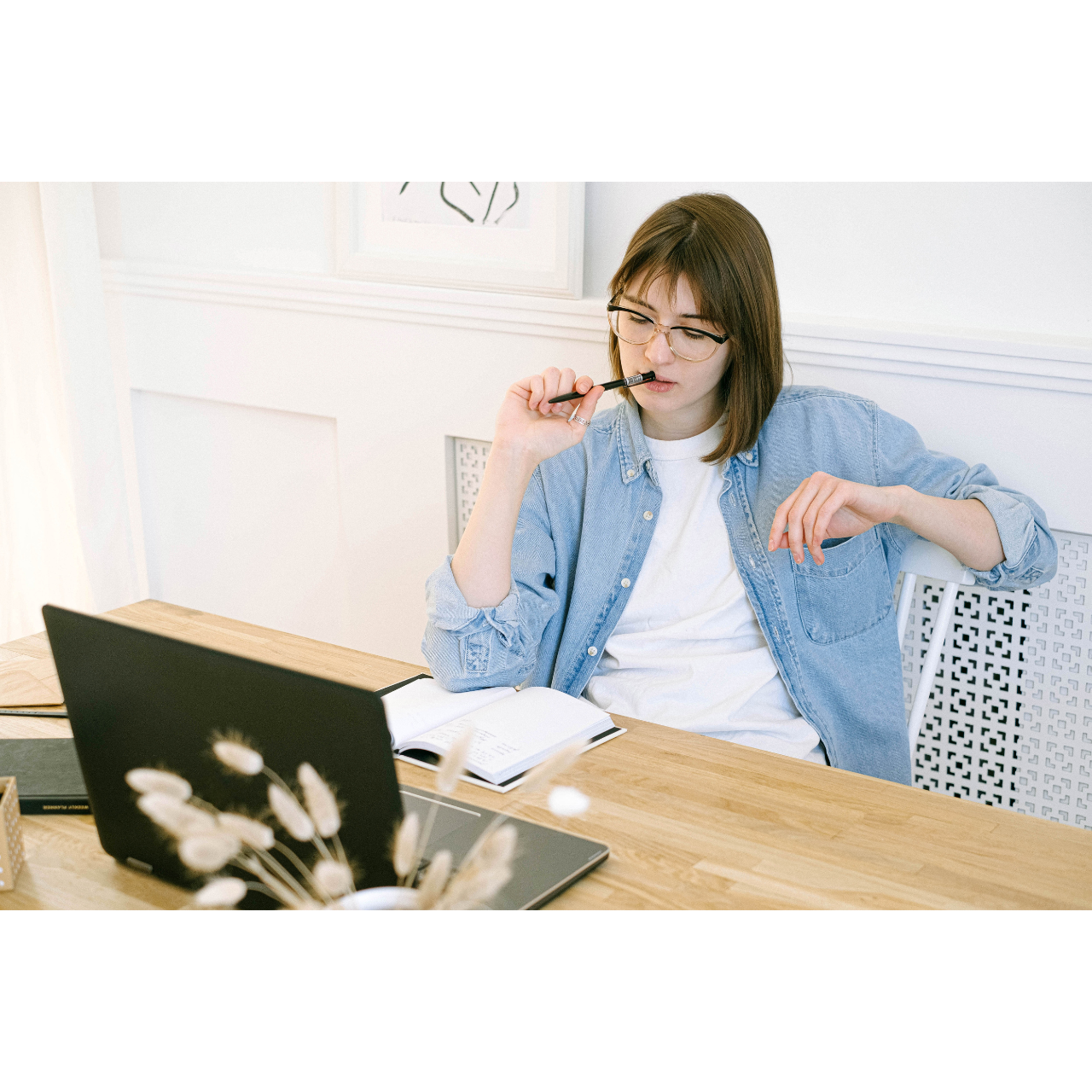 Person sitting at a wooden desk with a laptop, open notebook, and pen, in a bright home office setting with minimal decor.