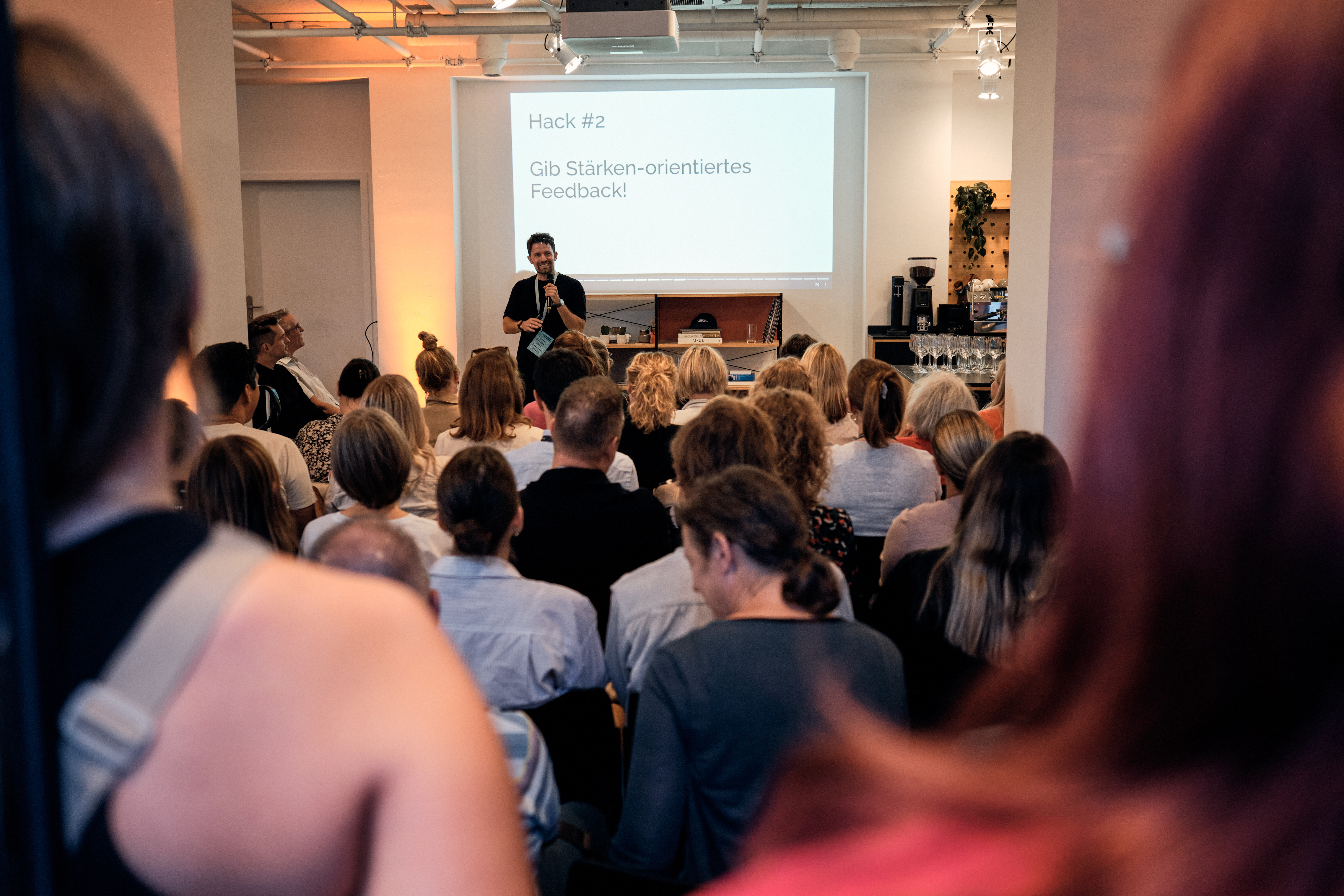 Eine Person hält vor einem sitzenden Publikum eine Präsentation, während auf eine Leinwand eine Folie mit der Aufschrift „Hack Nr. 2: Gib stärkeorientiertes Feedback!“ projiziert wird.