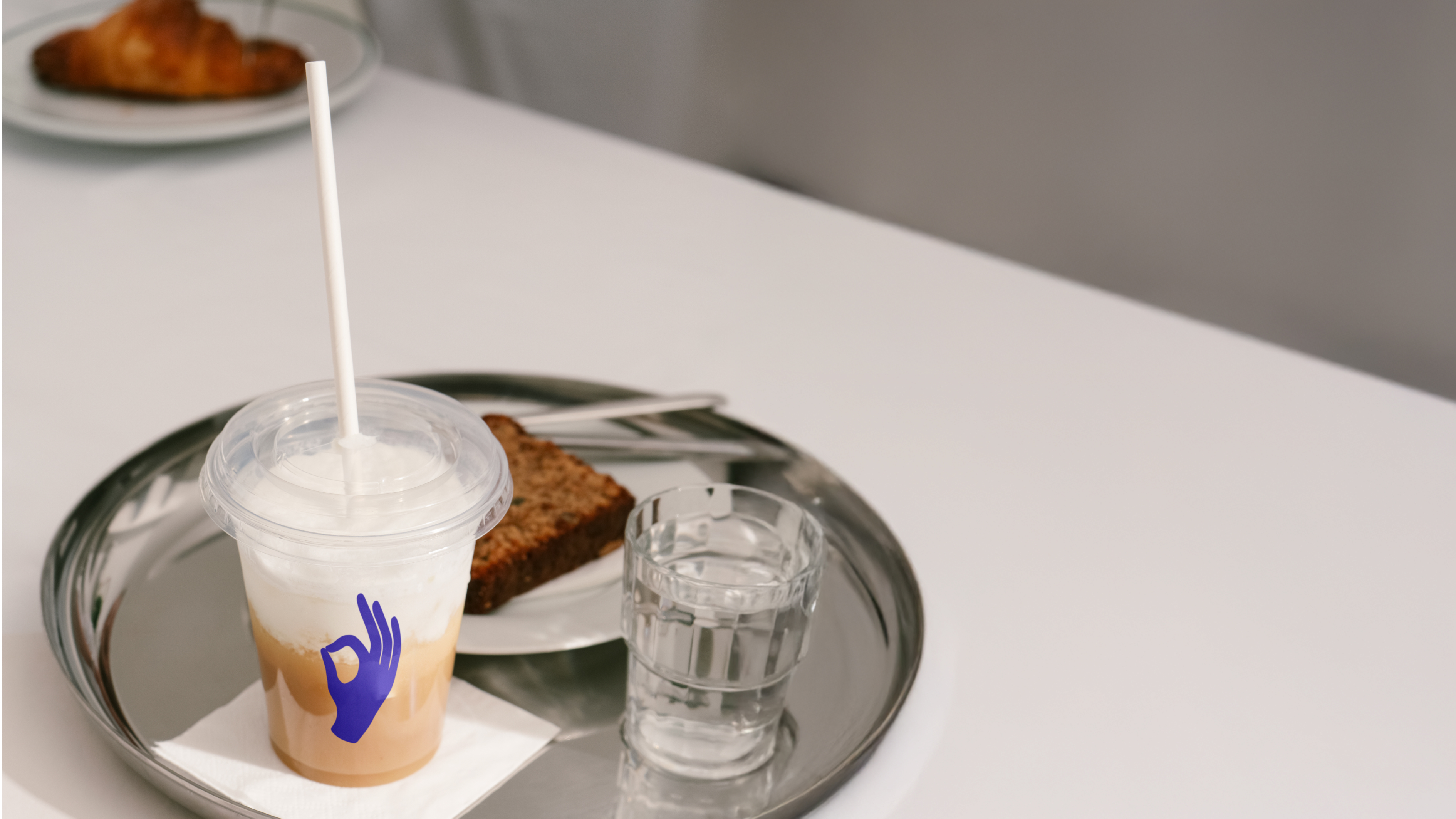 A plastic cup with iced coffee, a slice of bread, and a glass of water are served on a metal tray on a white table.