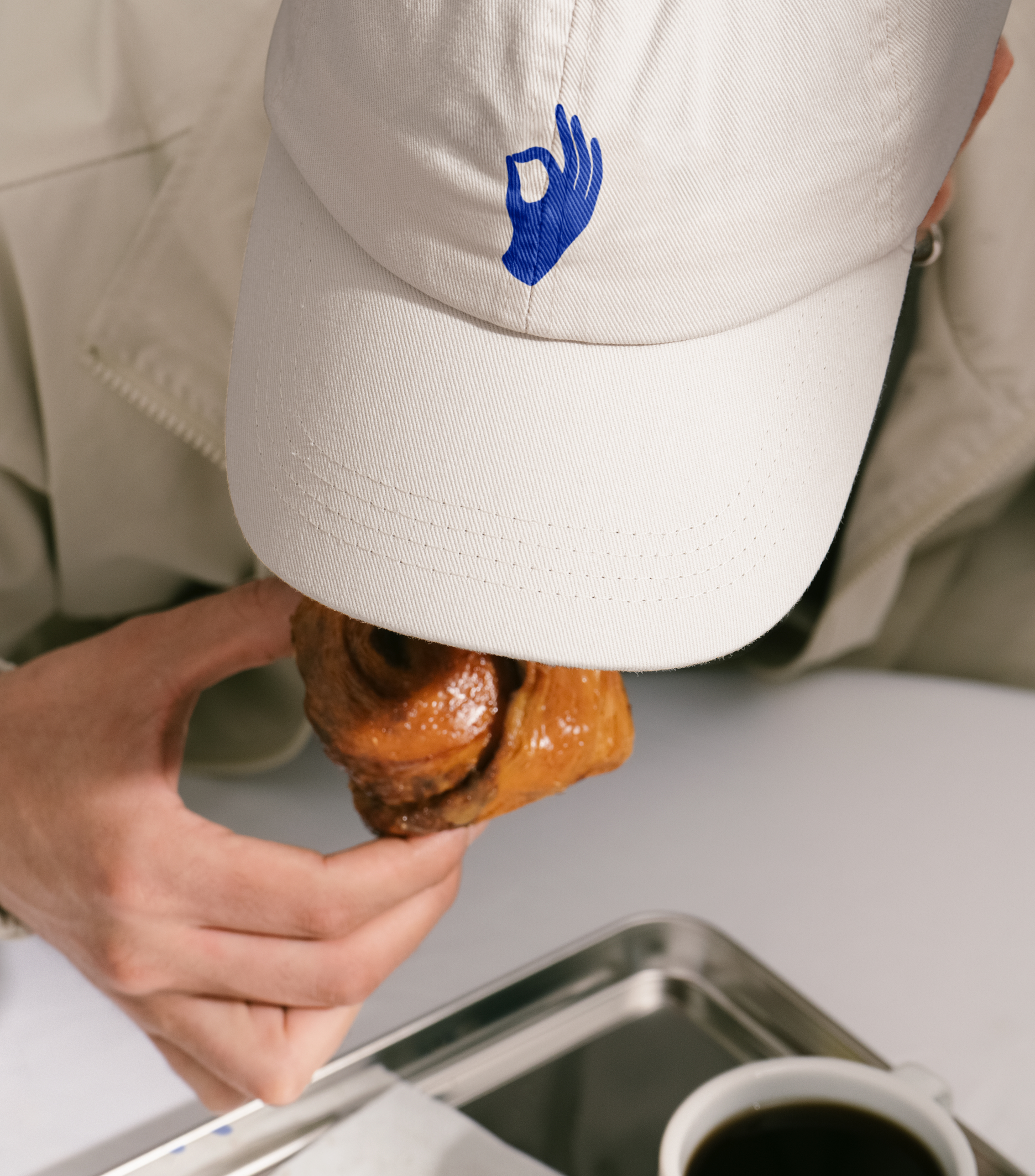 A person wearing a beige cap with a blue hand symbol takes a bite of a glossy cinnamon bun next to a cup of black coffee.