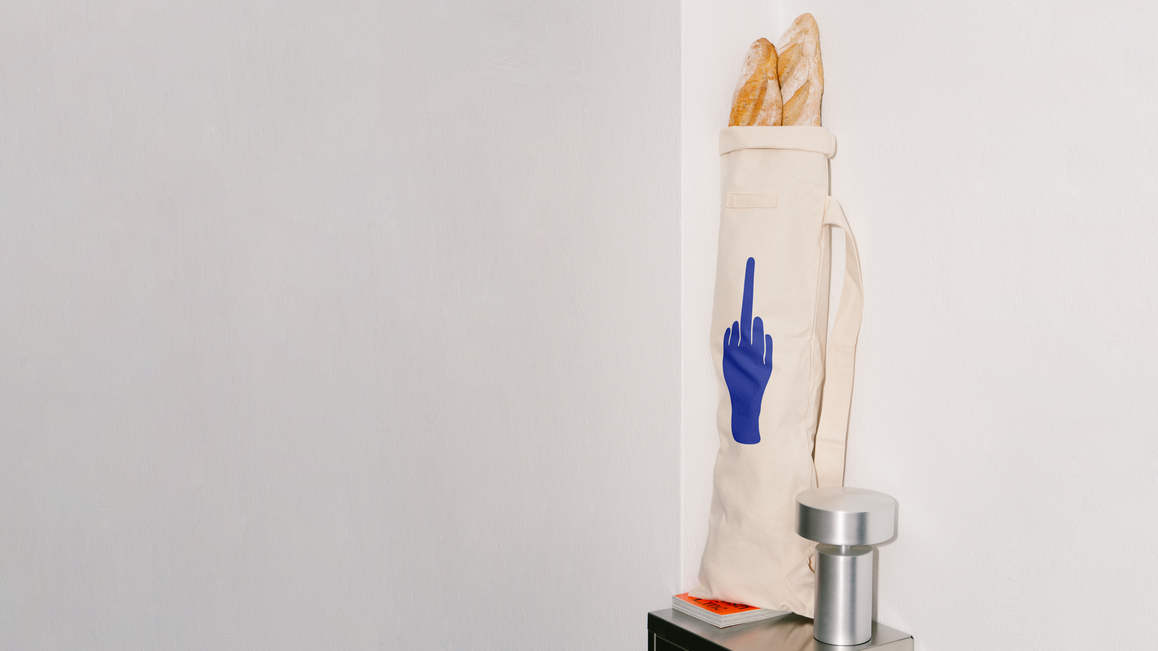 A cream-colored tote bag with a blue hand symbol showing the middle finger leans against a wall with baguettes sticking out.