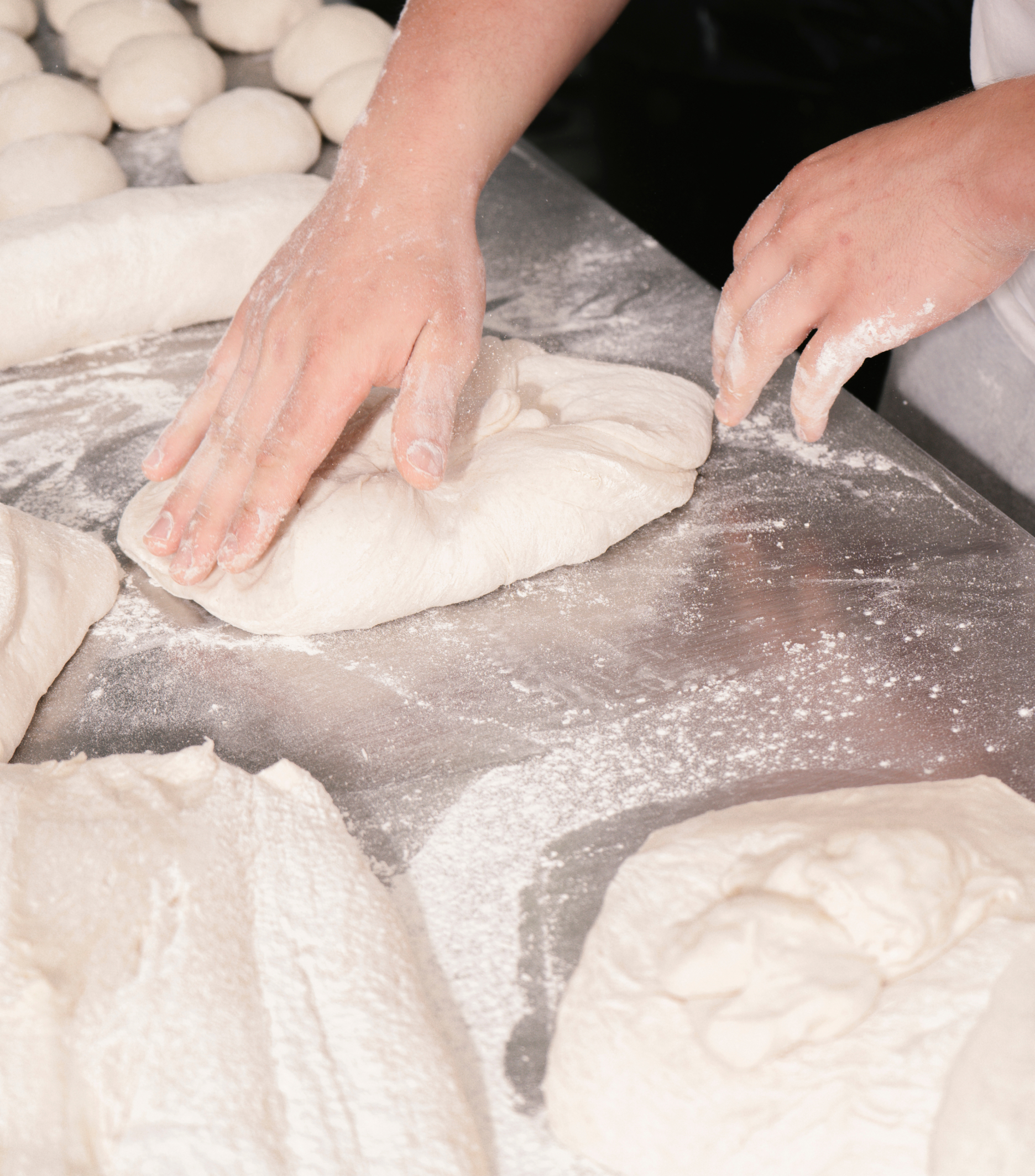 Two hands press and fold soft bread dough on a floured metal table surrounded by other dough pieces.