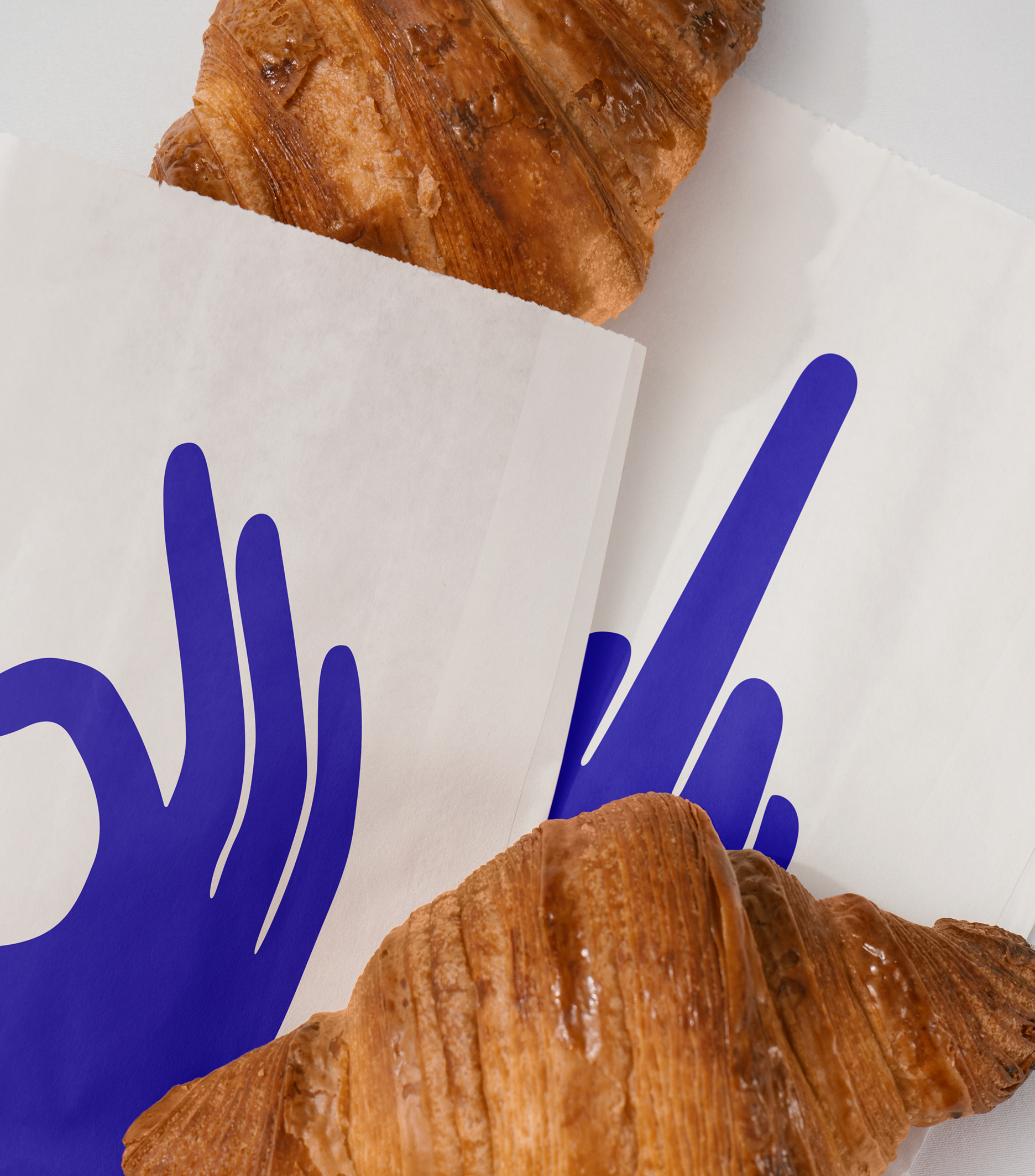 Two croissants peek out of paper bags printed with bold blue hand symbols showing different gestures.
