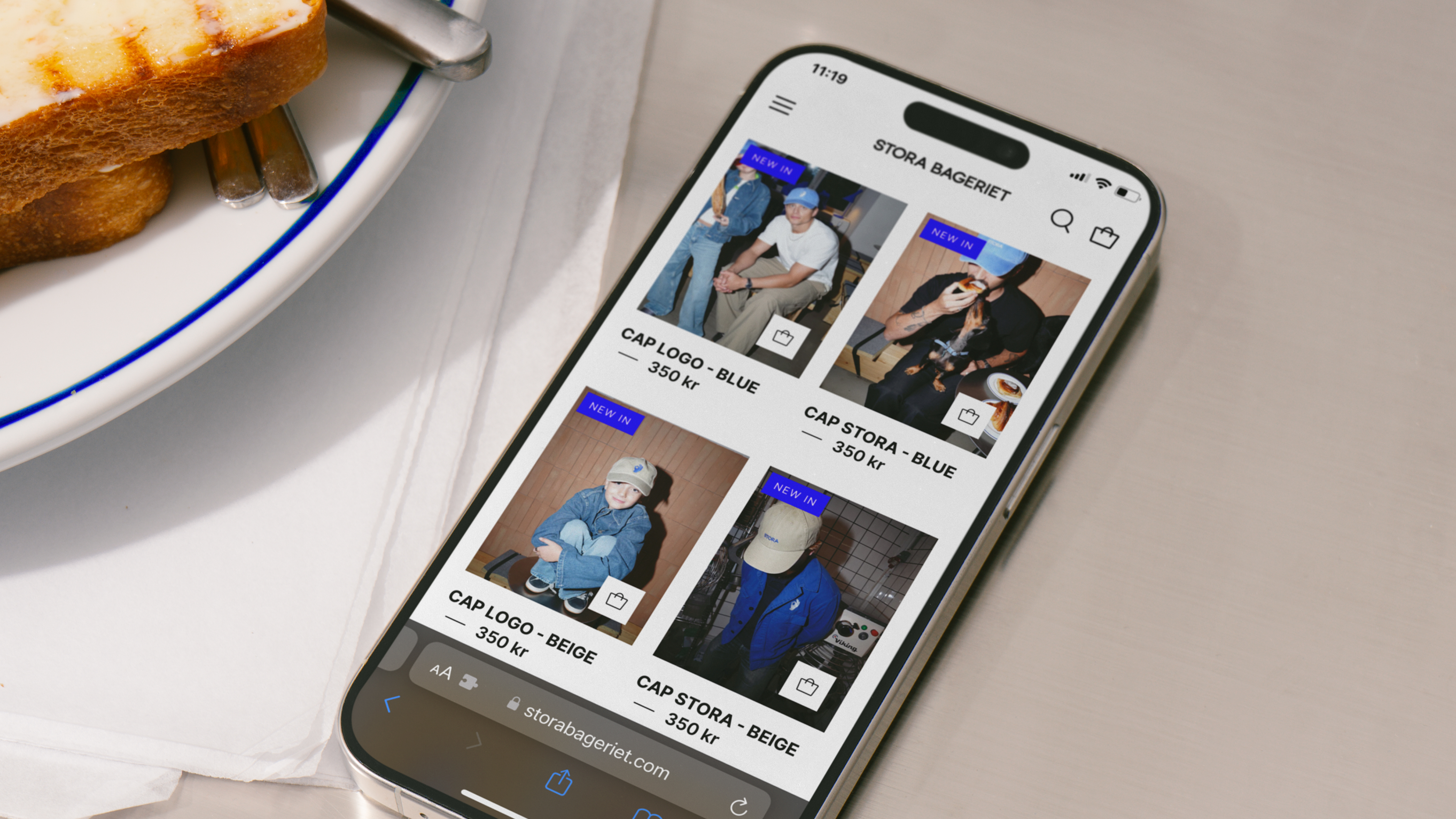 A smartphone displays Stora Bageriet’s webshop featuring branded caps, placed beside a plate with buttered toast.