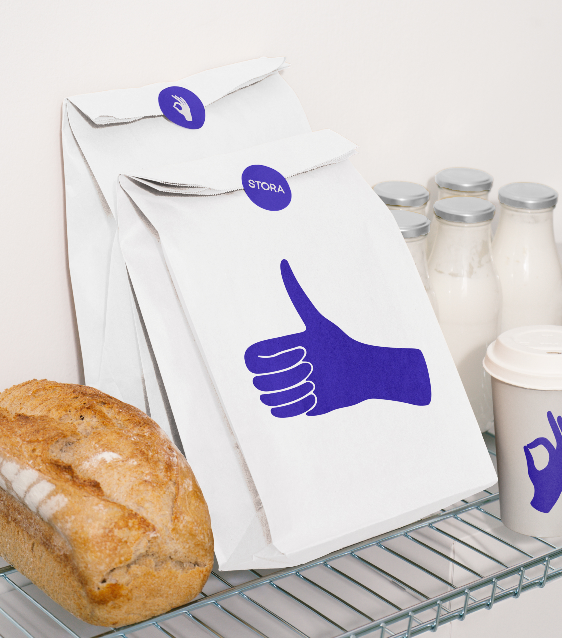 Paper bags with purple hand symbols and “Stora” stickers are displayed next to a loaf of bread, milk bottles, and a branded coffee cup.