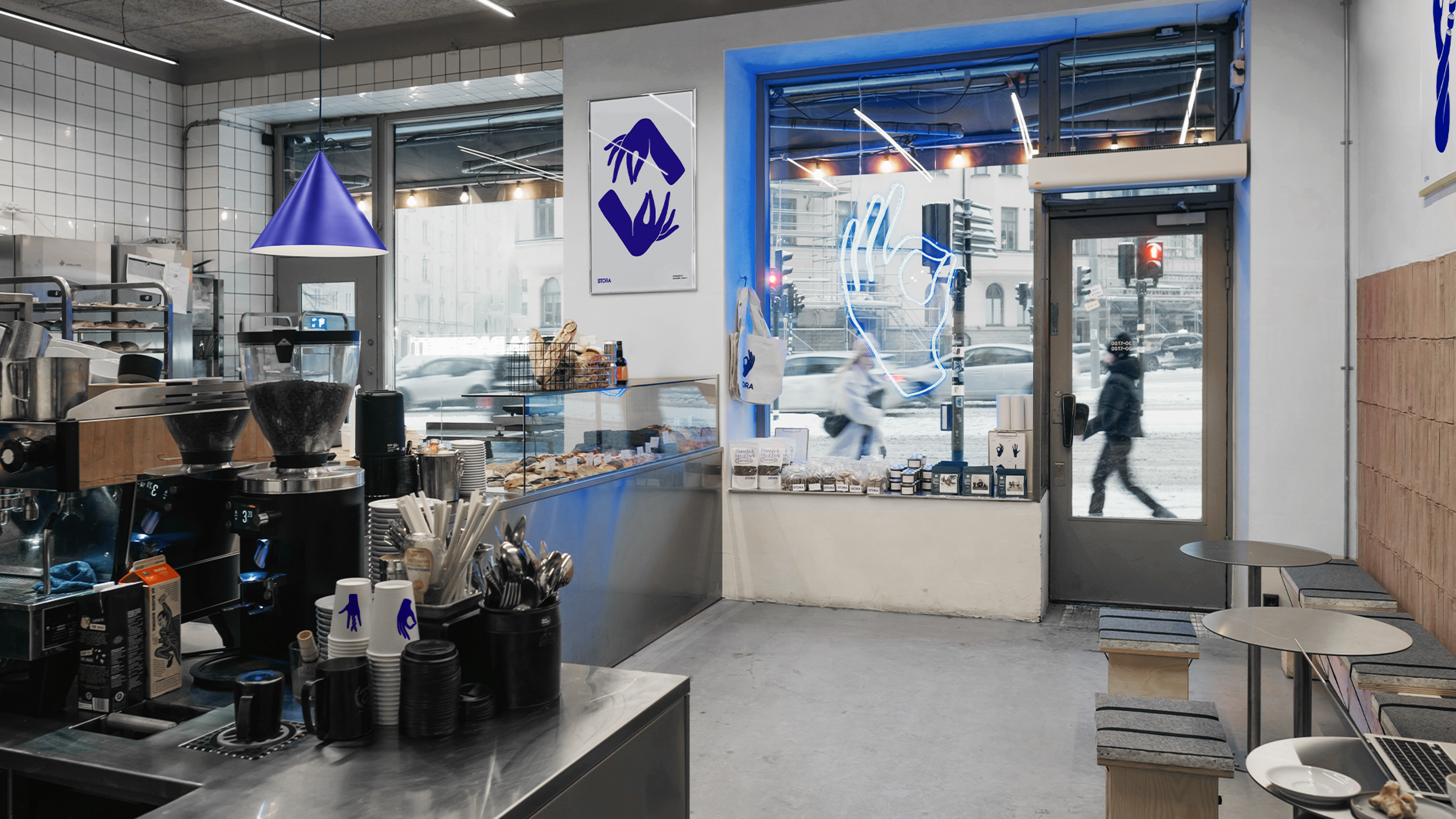 View of Stora Bageriet’s café interior with coffee machines, branded cups, baked goods, posters, and a neon hand sign in the window.
