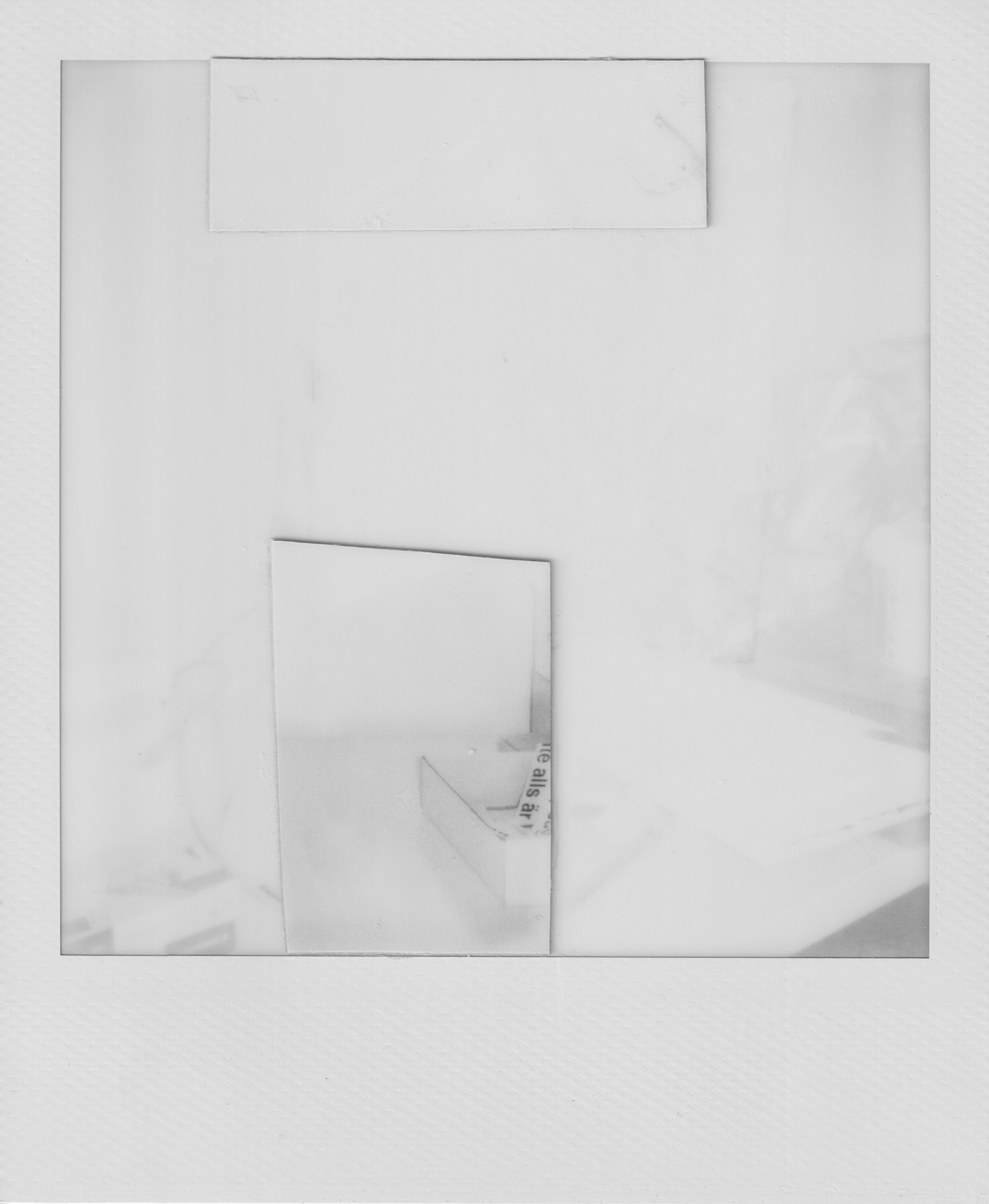 A scanned black and white Polaroid photograpgh of a small white paper box on a table, containing a cut-out paper stripe containing a text reading "te alls är".