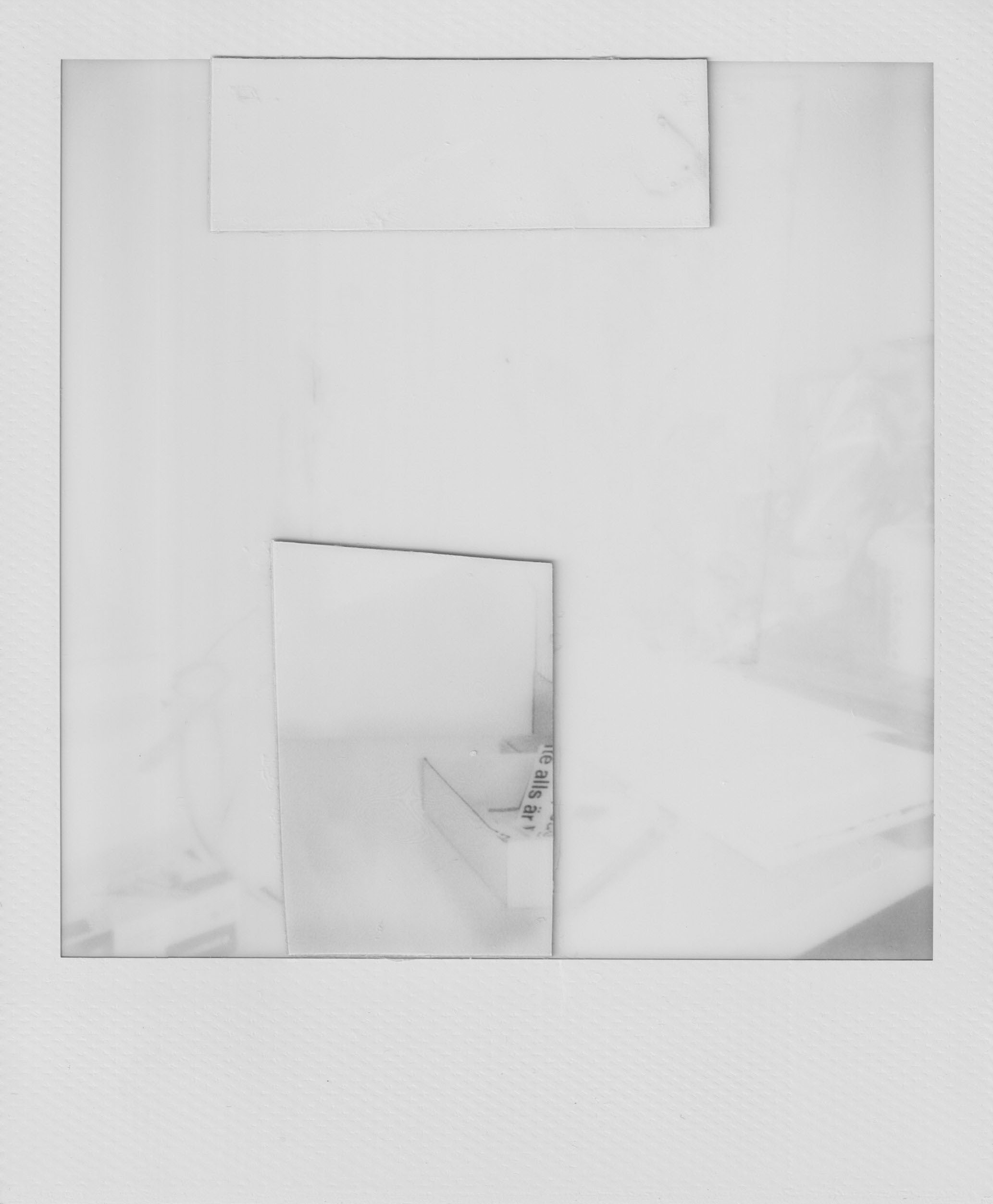 A highly overexposed black-and-white Polaroid photograph and abstract collage depicting a blurry table - although it’s hard to know for sure. Overlaid centrally are two roughly cut fragments of other Polaroid photographs: a horizontal strip across the top and a vertical rectangle near the center. The vertical fragment contains a box holding a cut-out paper strip bearing a snippet of printed text (“te allas är”) at its edge, hinting at a fragmented Swedish caption.