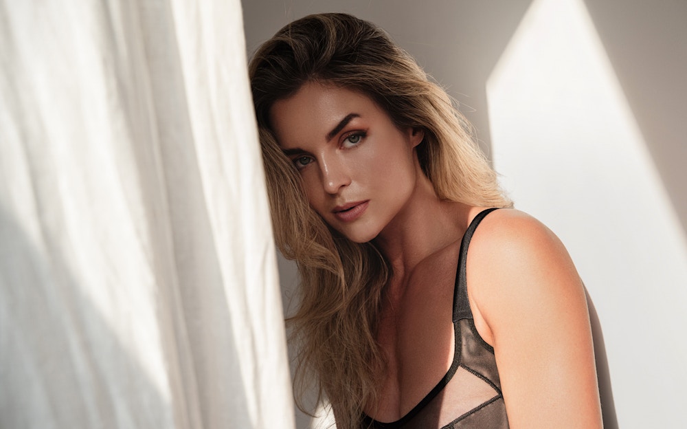 Woman seductively looks at the camera. A curtain blows a little to the let of her and her eyebrows are arched to show the seductive powers of a brow lift.