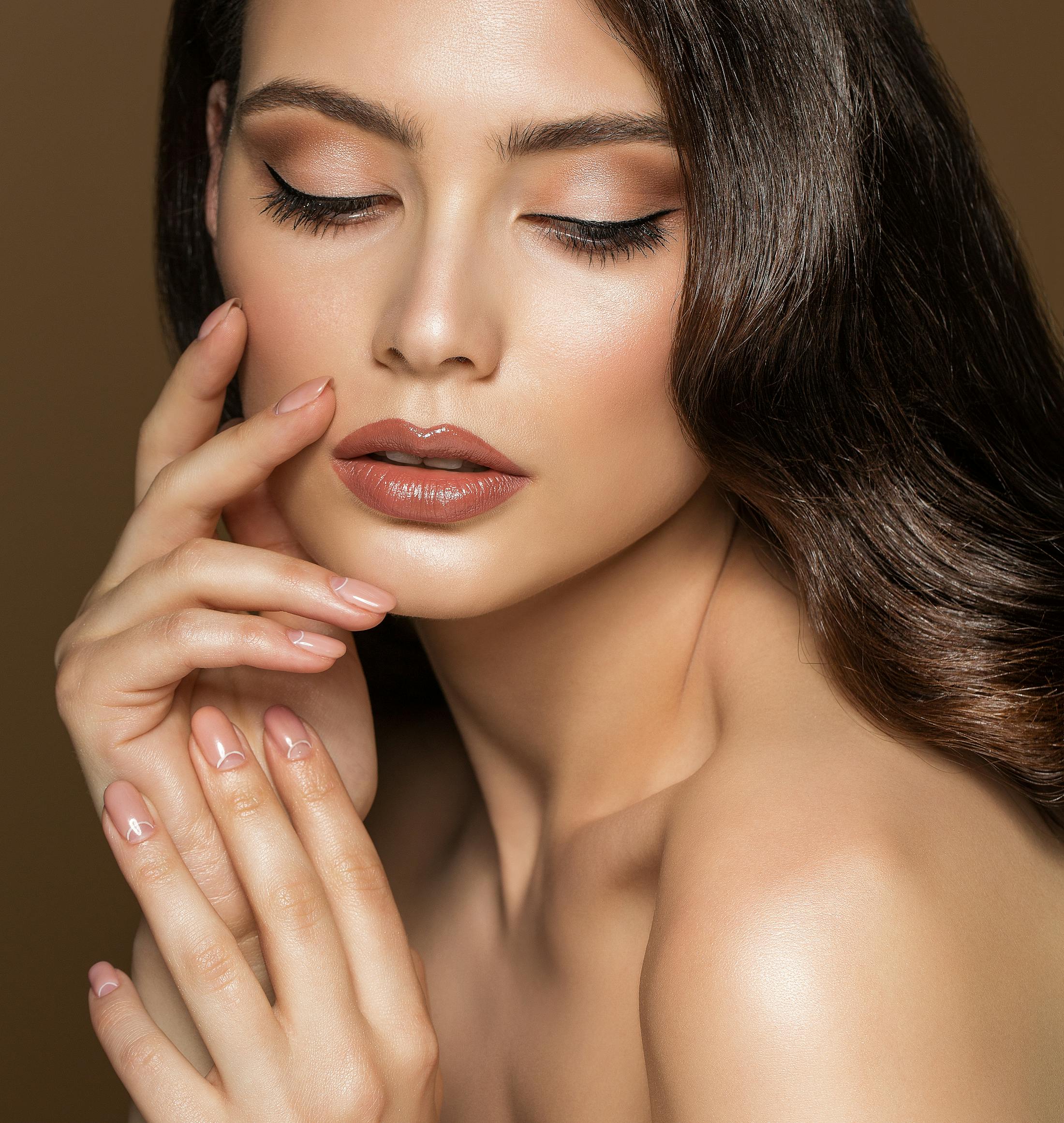 Stylizied portrait shot of a woman with full makeup and glowing skin. She looks to be shirless but the image is cropped abover her chest. Her hands are up by her face and one hand rests on her cheek. She had perfect eyebrows emulating a brow lift with Dr. Rahban in Beverly Hills