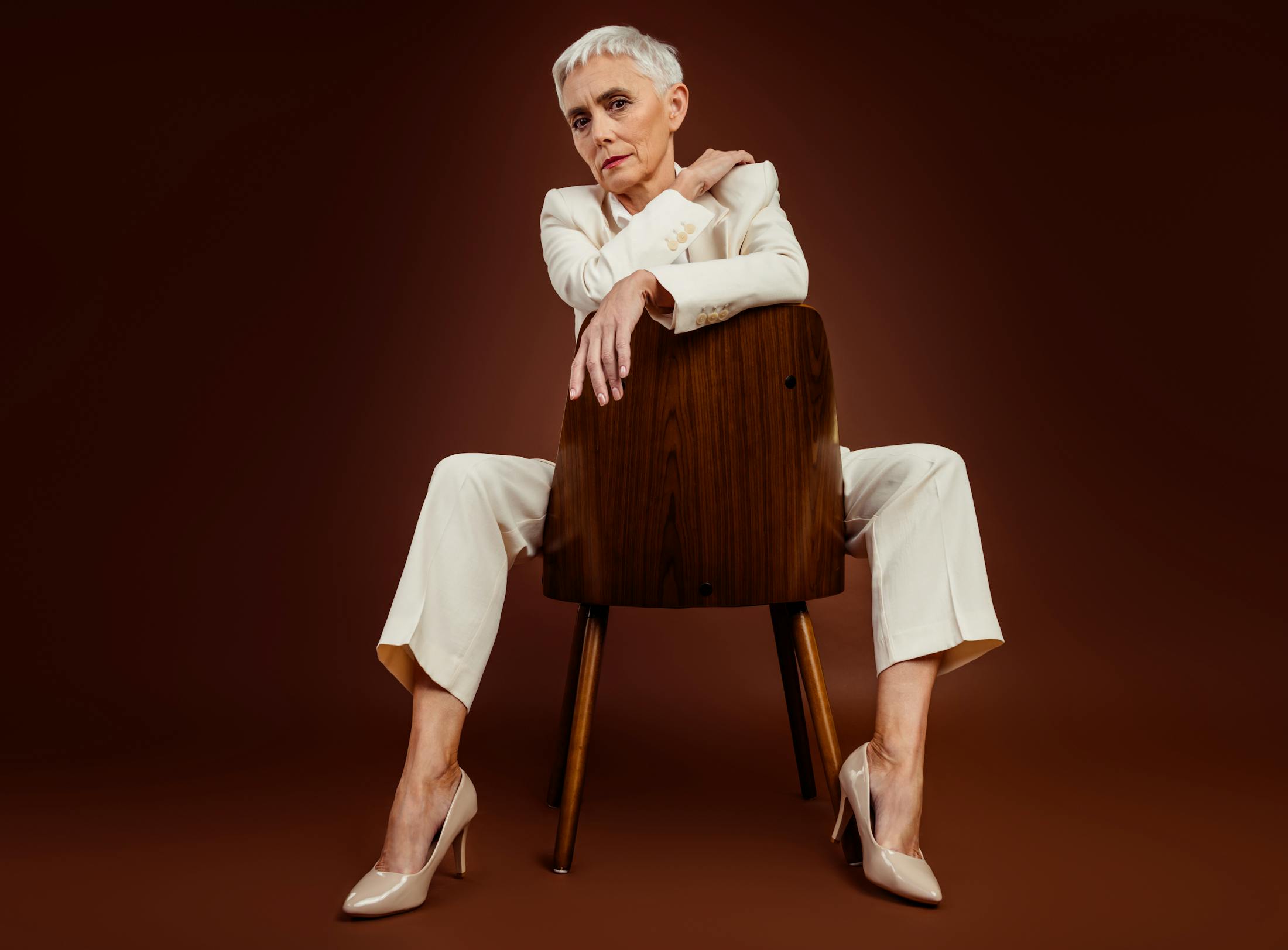 Woman in her 60's with a short gray pixie cute, straddles a mid-century chair while wearing a white suite and beige high heels. She looks confident, powerful, and stylish.
