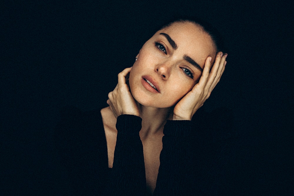 Woman in her 40's with a black long sleeve shirt on poses effortlessly at the camera with her elbows rested her hands are able to be around her neck and face. She has a stunning face with no wrinkles to show the potential results of a Beverly Hills Mini Facelift.