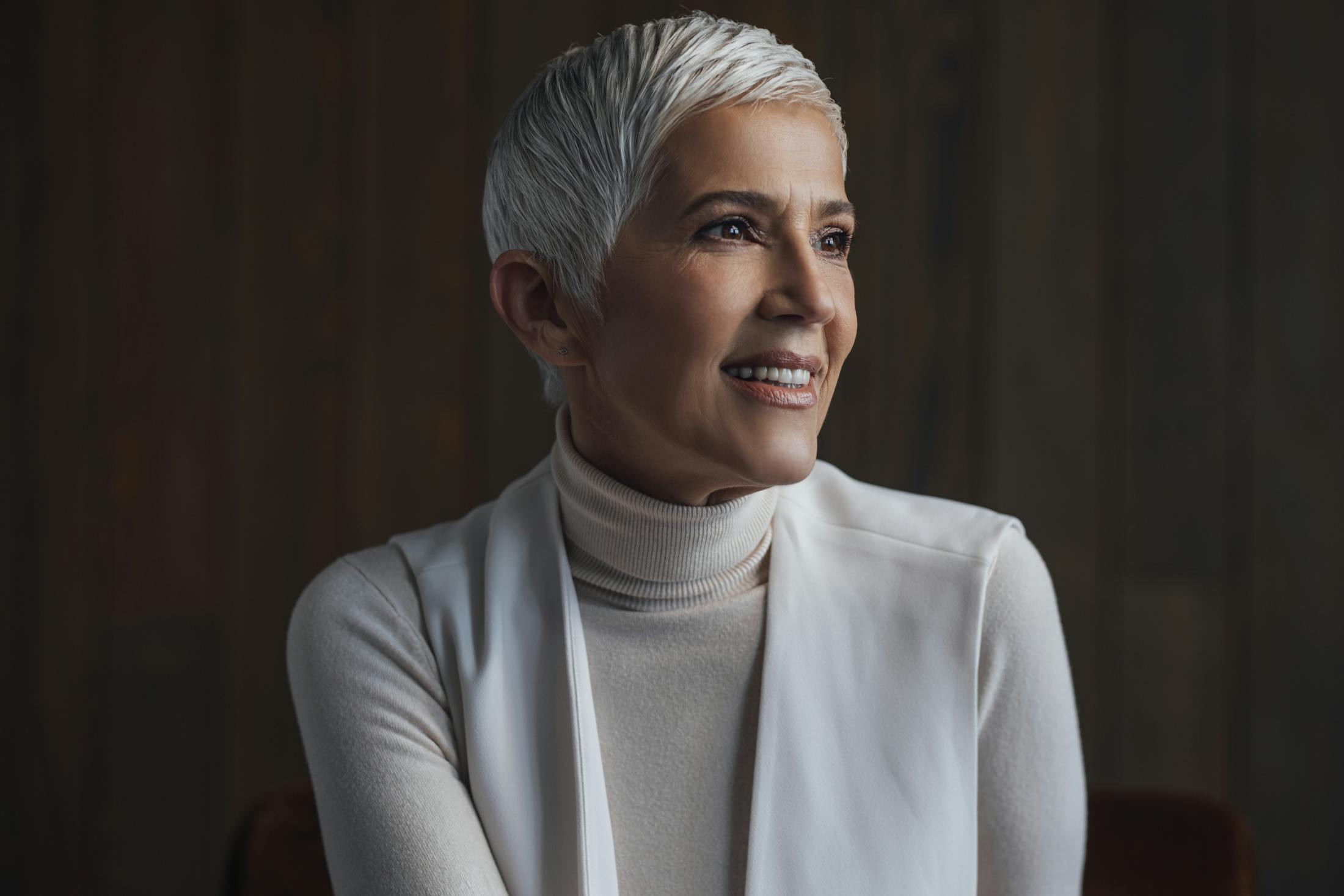 Woman in her late 50's with a gray pixie cut looks off to the side while smiling. She is wearing a white turtleneck with a white vest. The background is dark to show a nice contrast and the womans youthful appearance despite her age is a good example of the potential with a Beverly Hills Facelift by Dr. Rahban