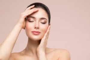 iStock 1301188932Beautiful Woman Face Skin Care. Natural Make up. Skin Health Treatment. Model massaging Face by Hands. Beige Studio background. Fashion Girl with Closed eyes